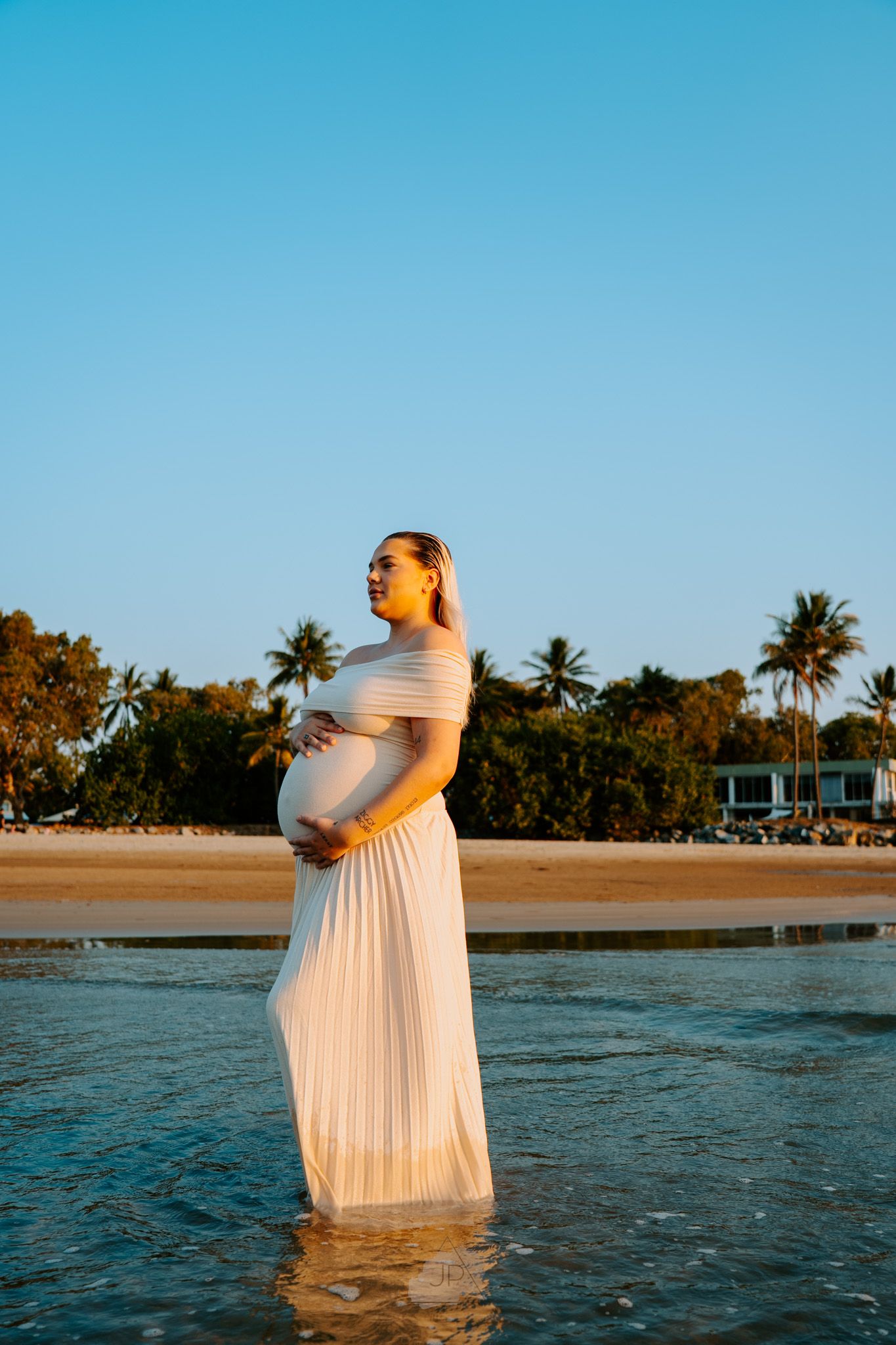 Woman in white dress stands in ocean water at sunset with palm trees and beach houses in background.