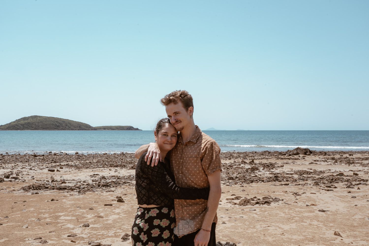 A couple embraces on a rocky beach with ocean waves and blue sky in the background.