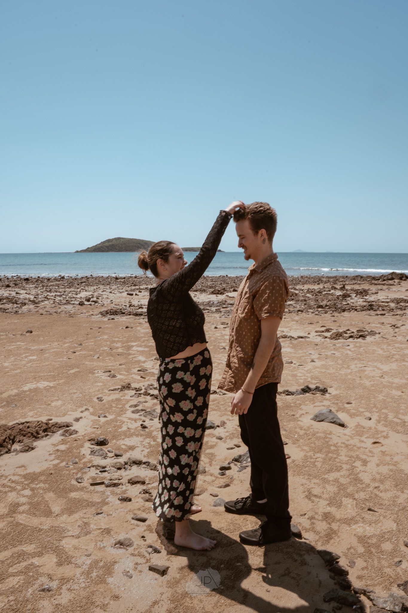 A couple shares a playful moment on a rocky beach with an island visible in the distance on a clear day.