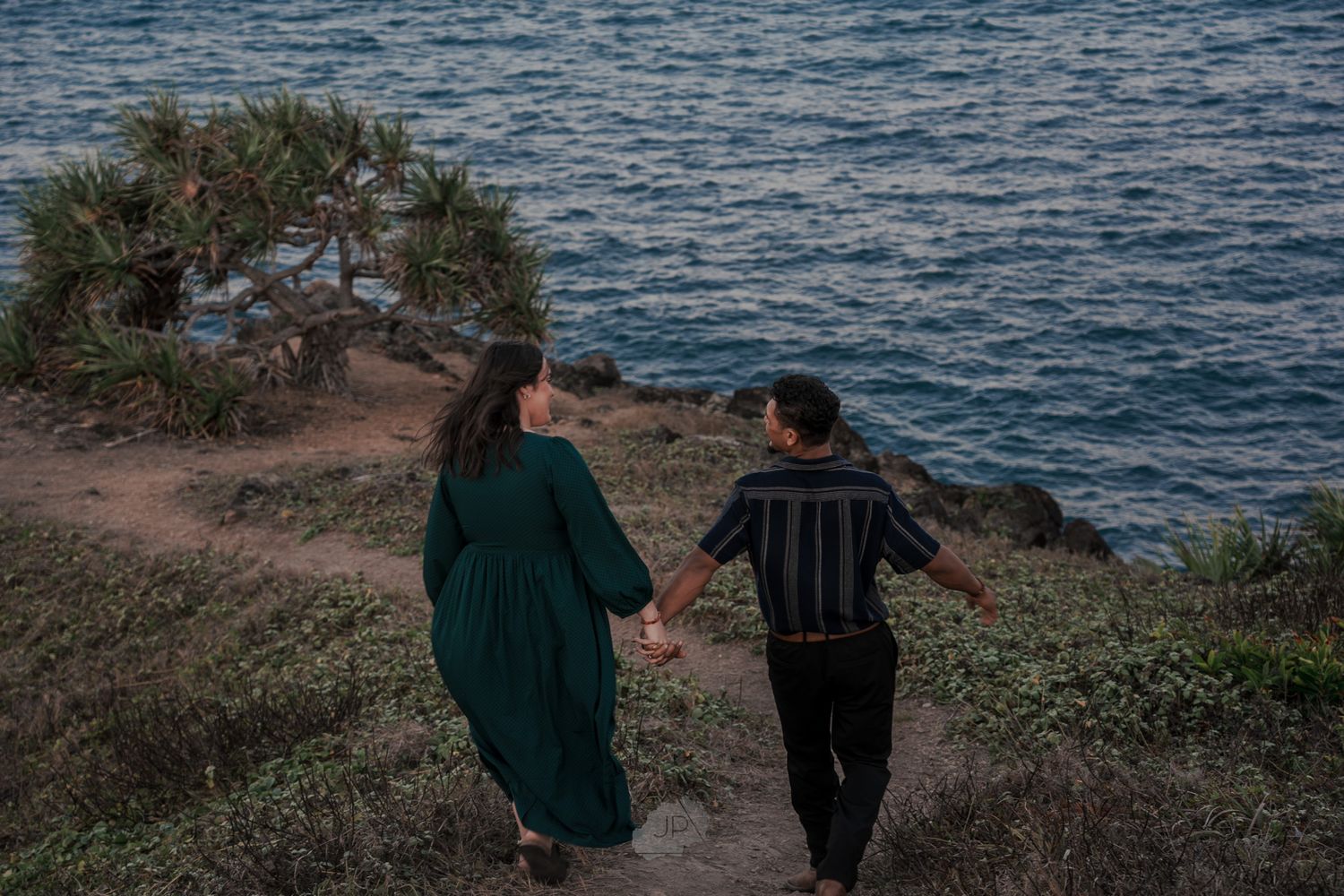 A couple walks along a scenic coastal cliff path overlooking the blue ocean at sunset.