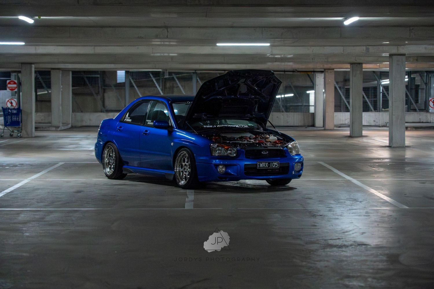 A blue Nissan Skyline GTR is parked in a dimly lit concrete parking garage.