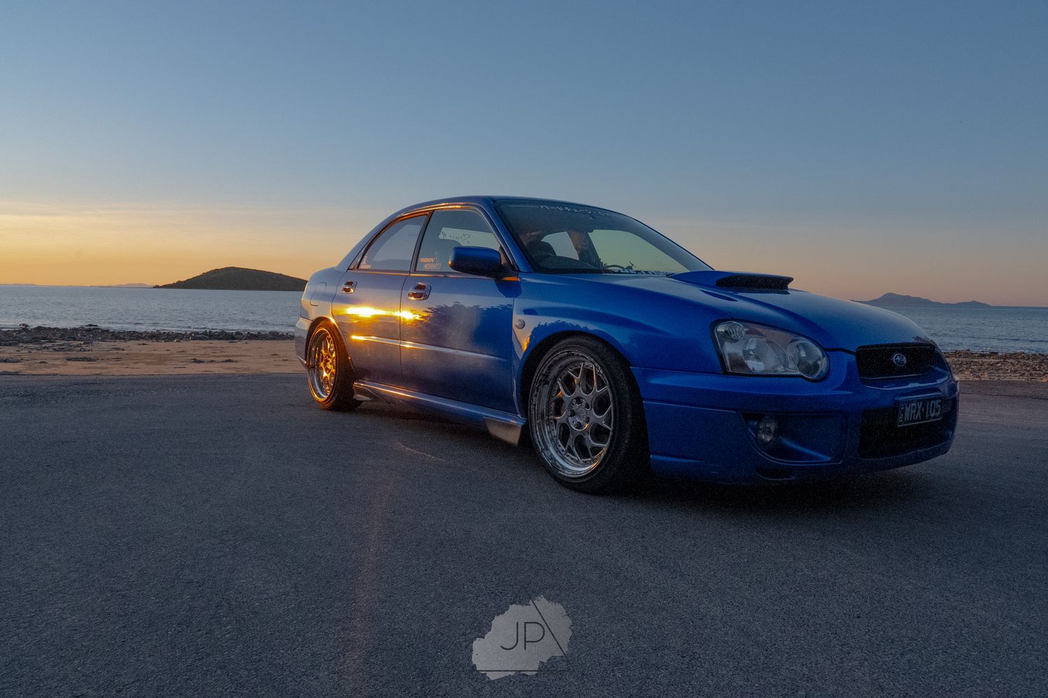 Blue Subaru WRX STI sports car parked on beach during sunset with dramatic lighting and coastal scenery.