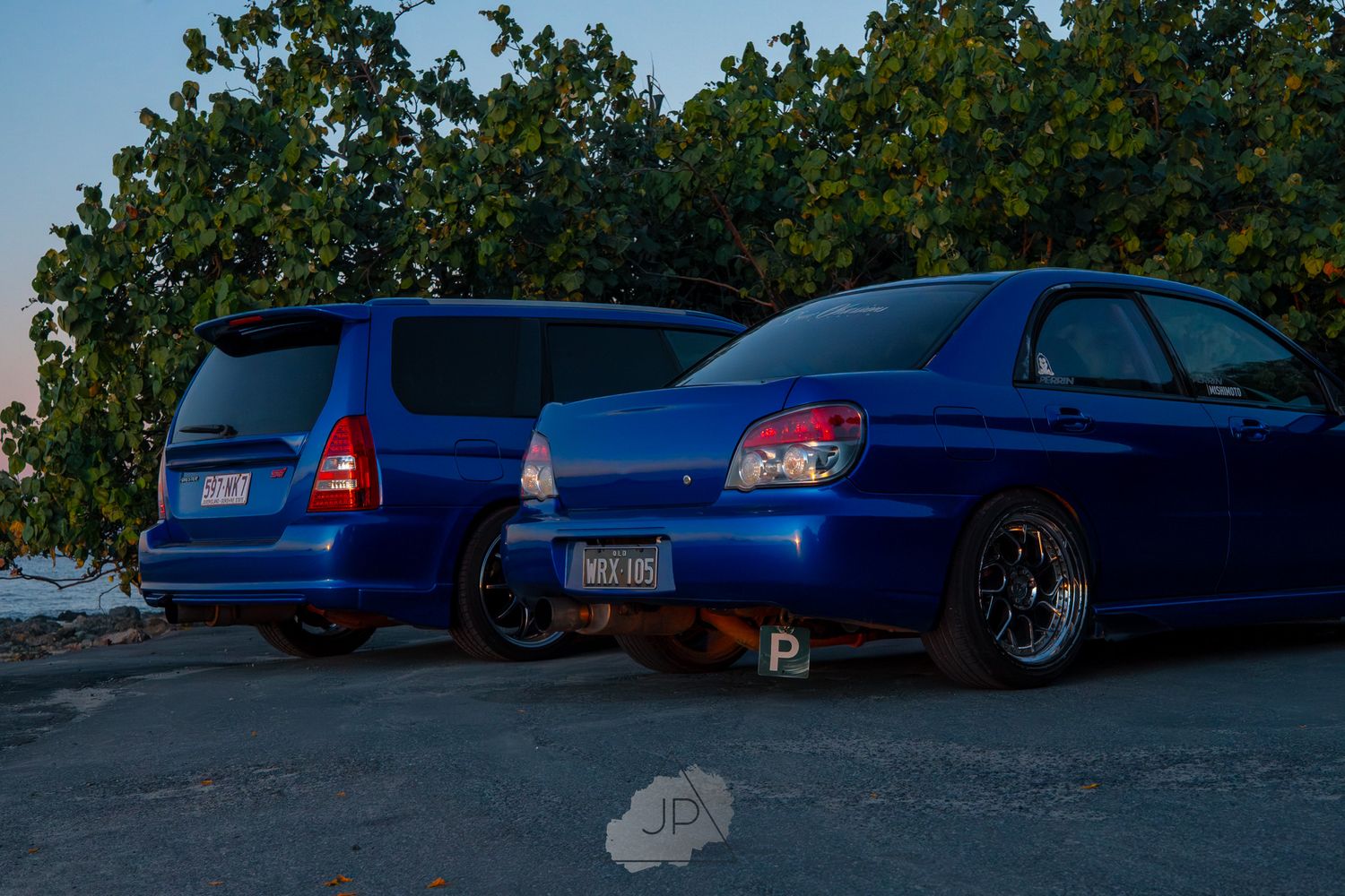 Blue sedan cars parked in a row at sunset with trees visible in the background providing shade.
