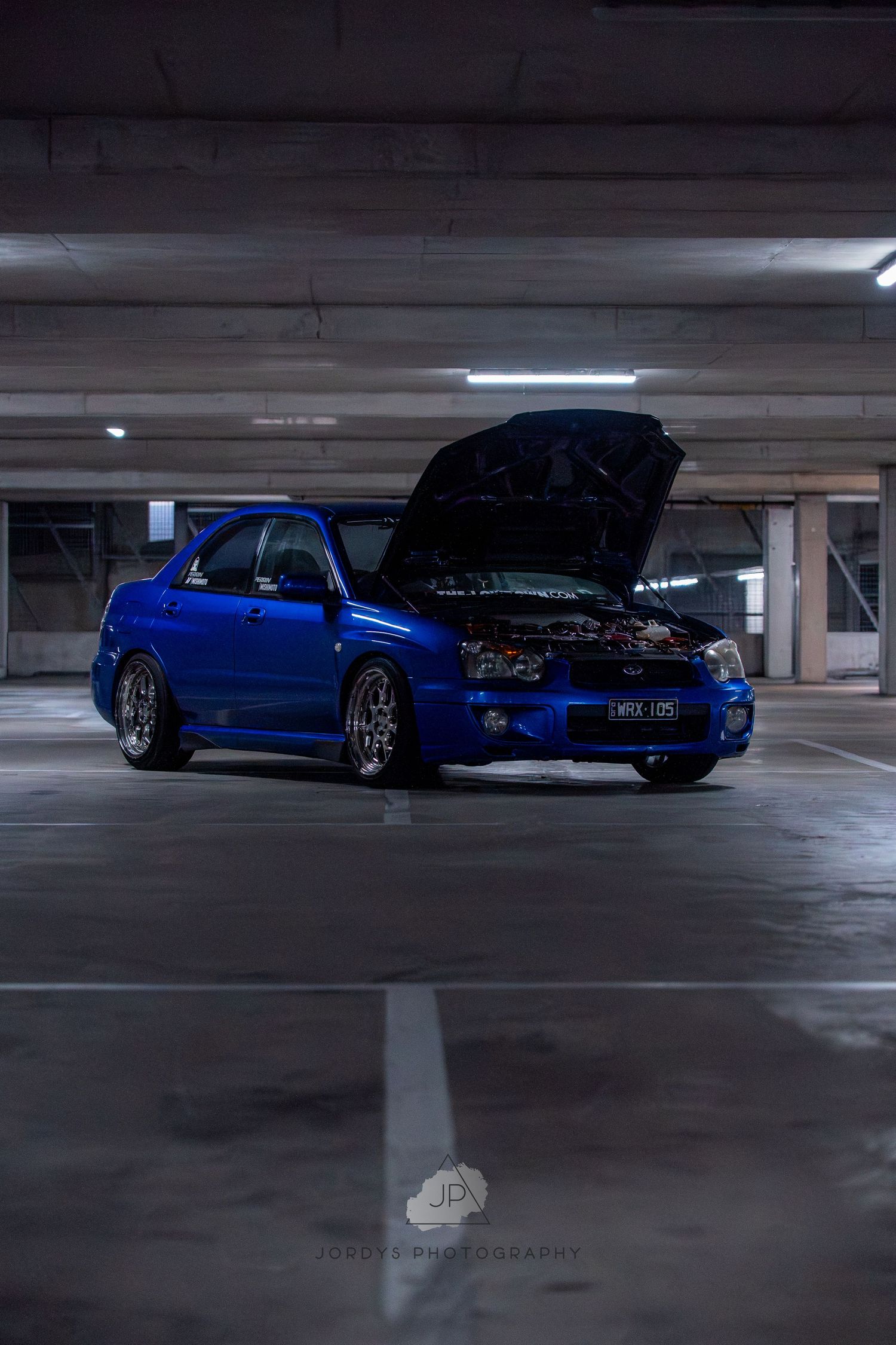 A modified blue Subaru WRX STI with its hood open sits in a dimly lit parking garage.