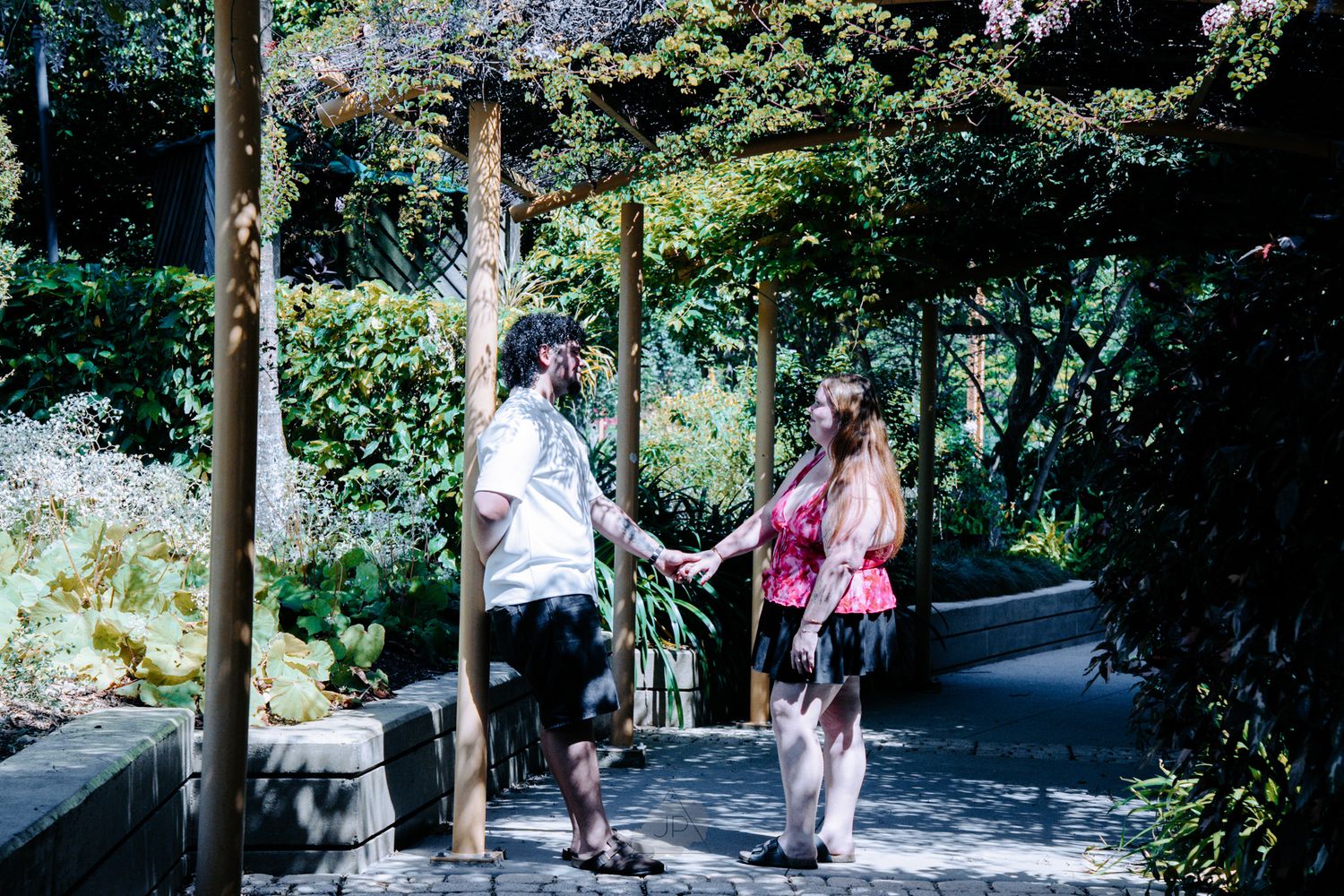 People walking along a winding garden path surrounded by lush greenery and trees on a sunny day.