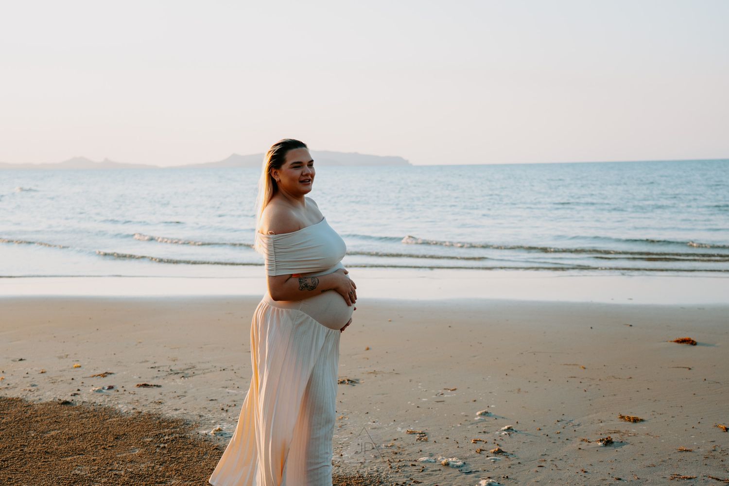 A romantic couple in white clothing embraces on a sandy beach at sunset with ocean waves in the background.