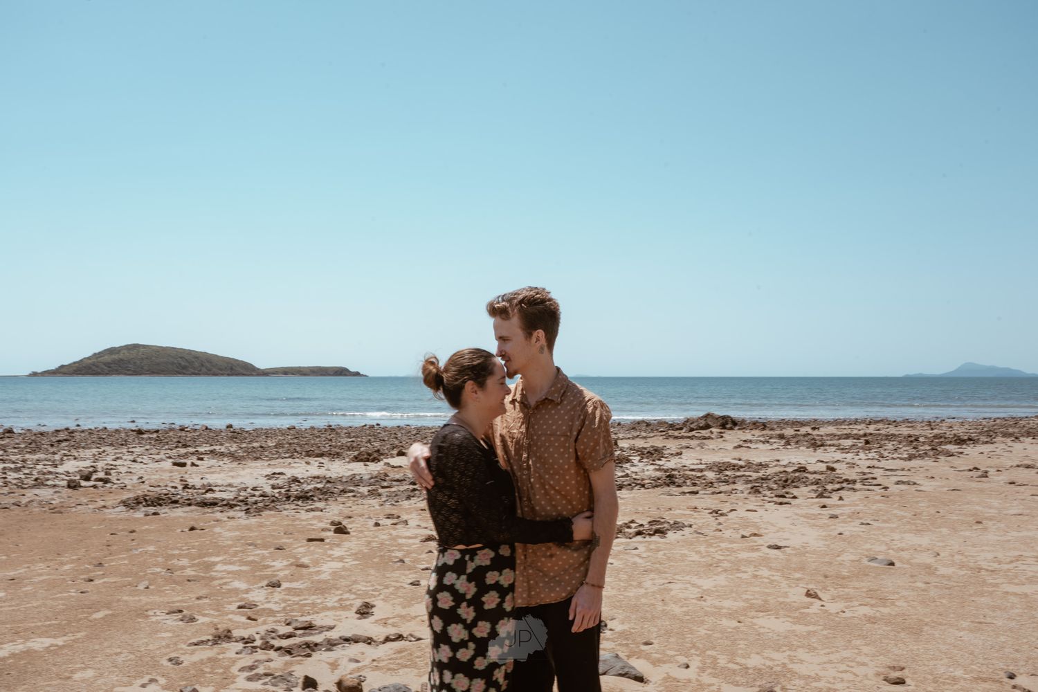A couple embraces on a rocky beach shoreline with islands visible in the distance under blue skies.