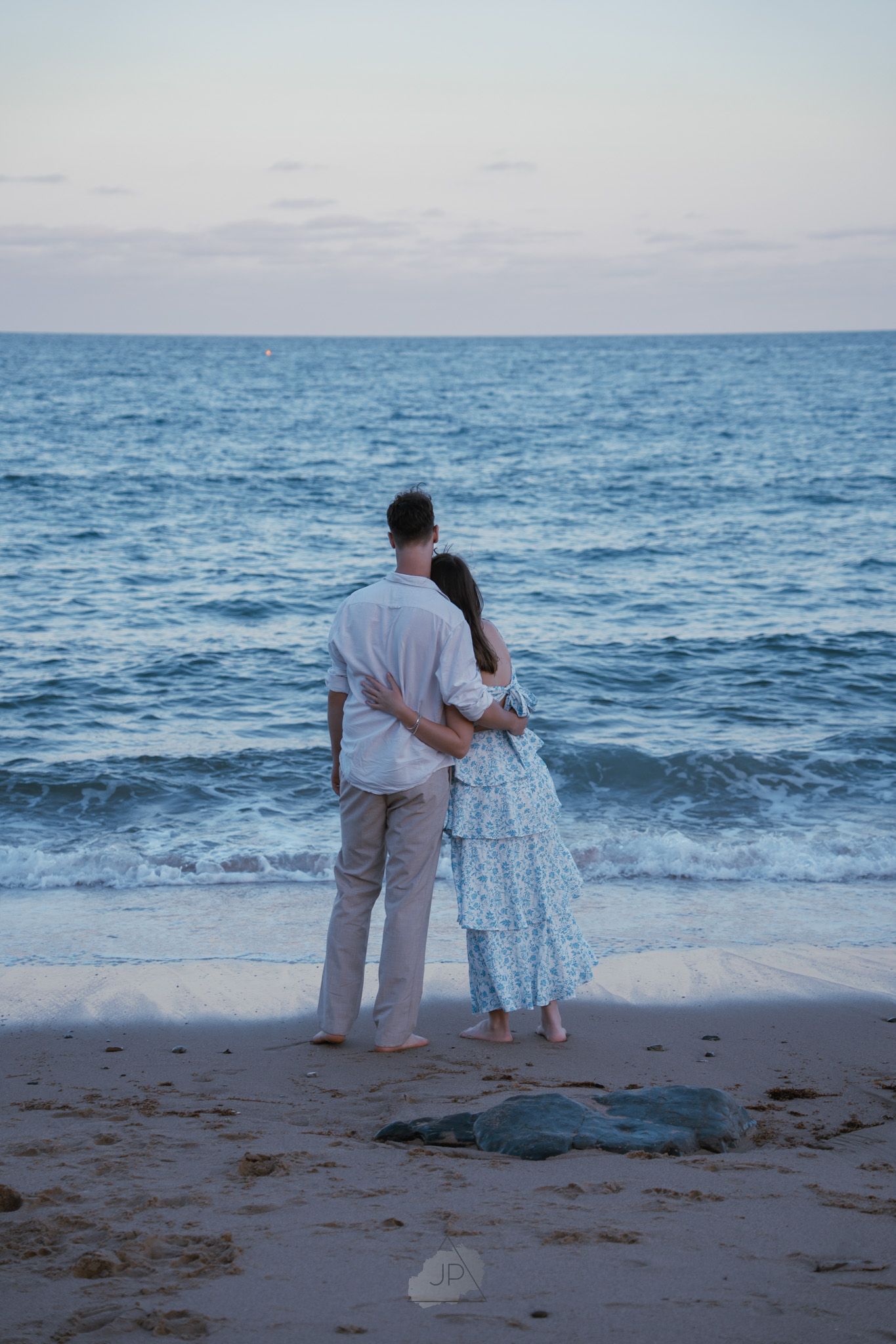Two people observe a sea turtle on a sandy beach at sunset with ocean waves in the background.