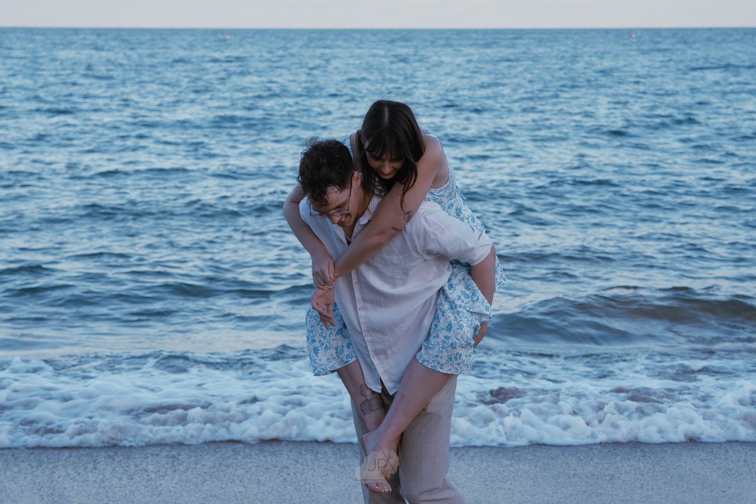 A couple embraces on the beach with the blue ocean waves rolling in during sunset.