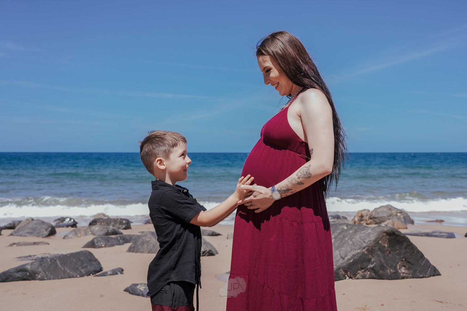 A series of beach photos showing tender moments between a pregnant woman in burgundy dress and young child on a rocky shore.