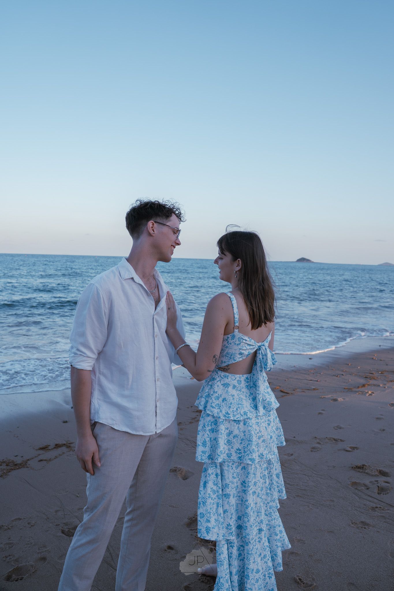 A couple stands together on a sandy beach with ocean waves and blue sky in the background.