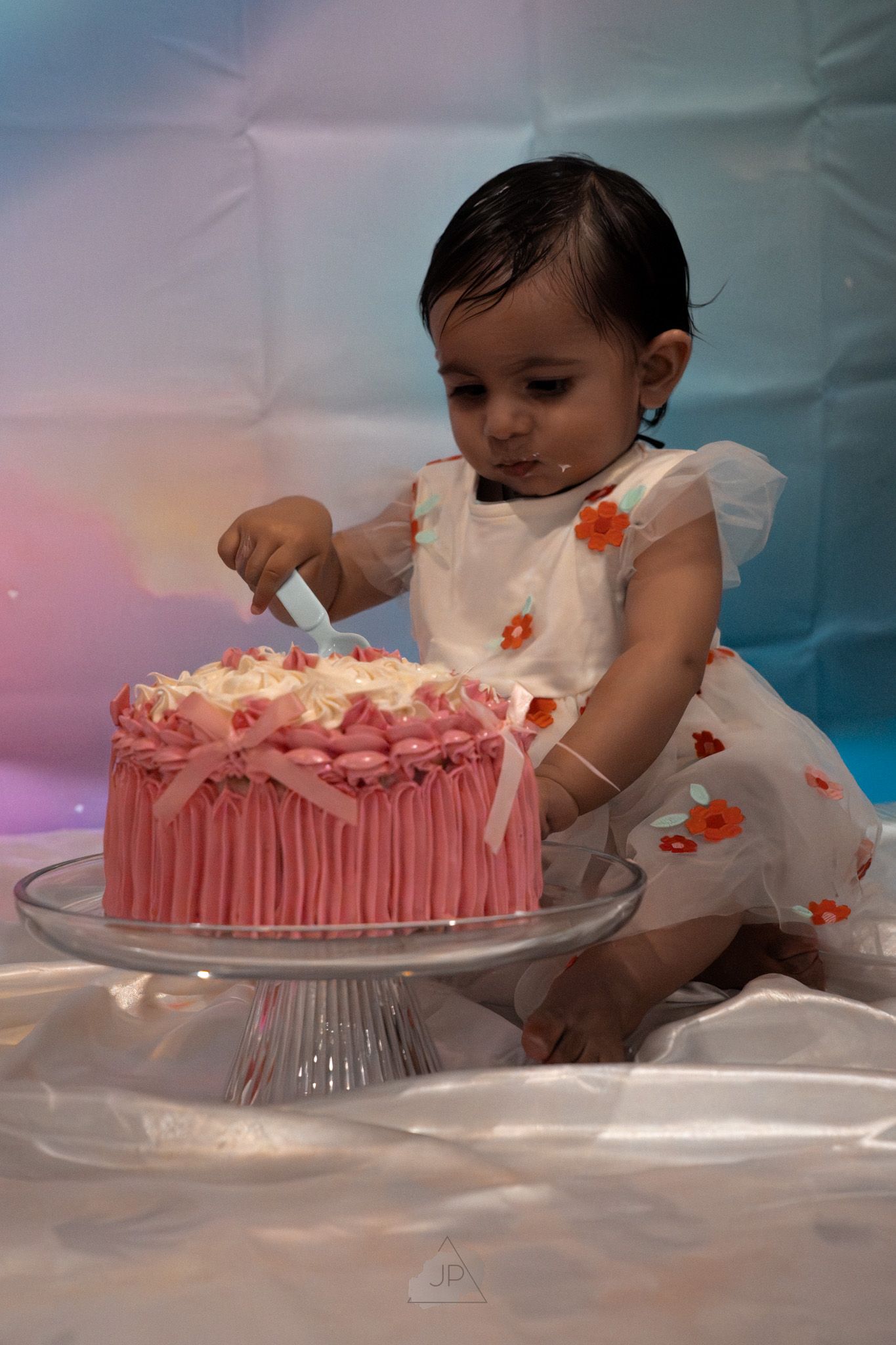 A beautifully decorated pink birthday cake sits on a glass cake stand with delicate floral decorations.