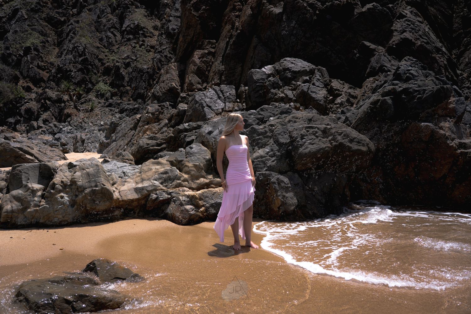 A person in a pink dress walks along a sandy beach near dark rocky cliffs and crashing waves.