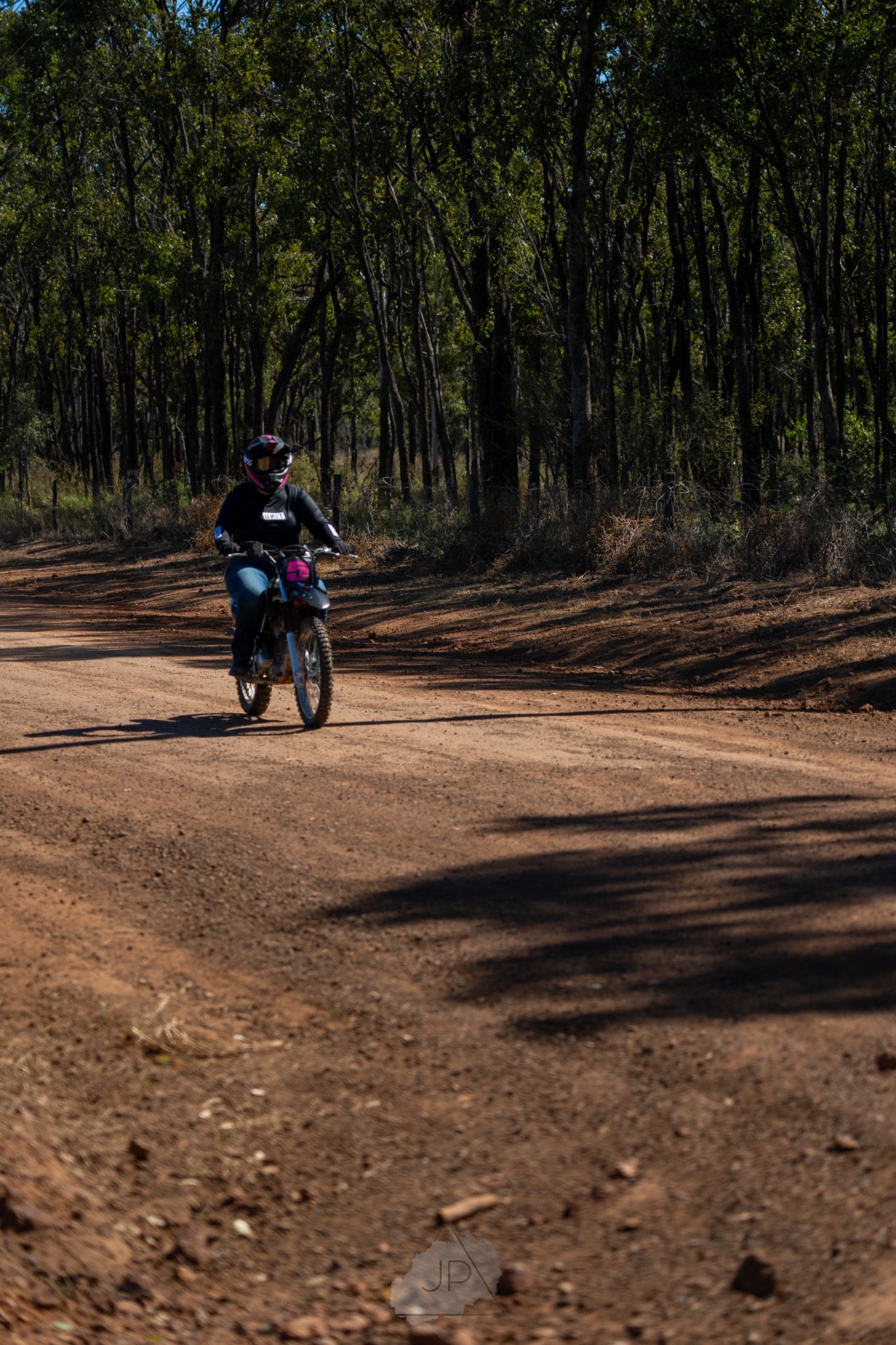 A dirt bike rider navigates through a wooded forest trail on a sunny day, casting long shadows on the dusty ground.