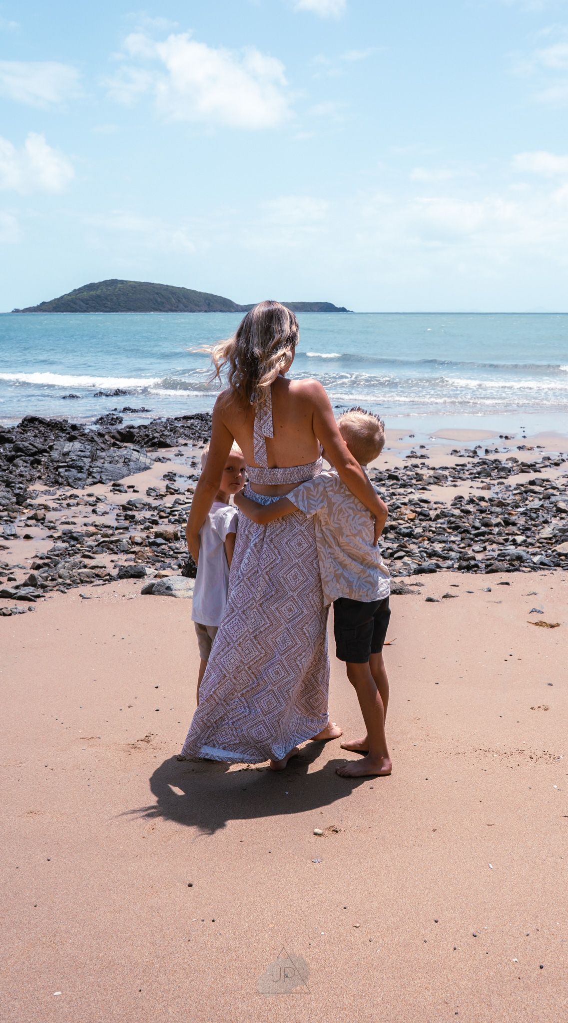 Two people sitting together on a rocky beach looking out at the ocean and distant island on a sunny day.