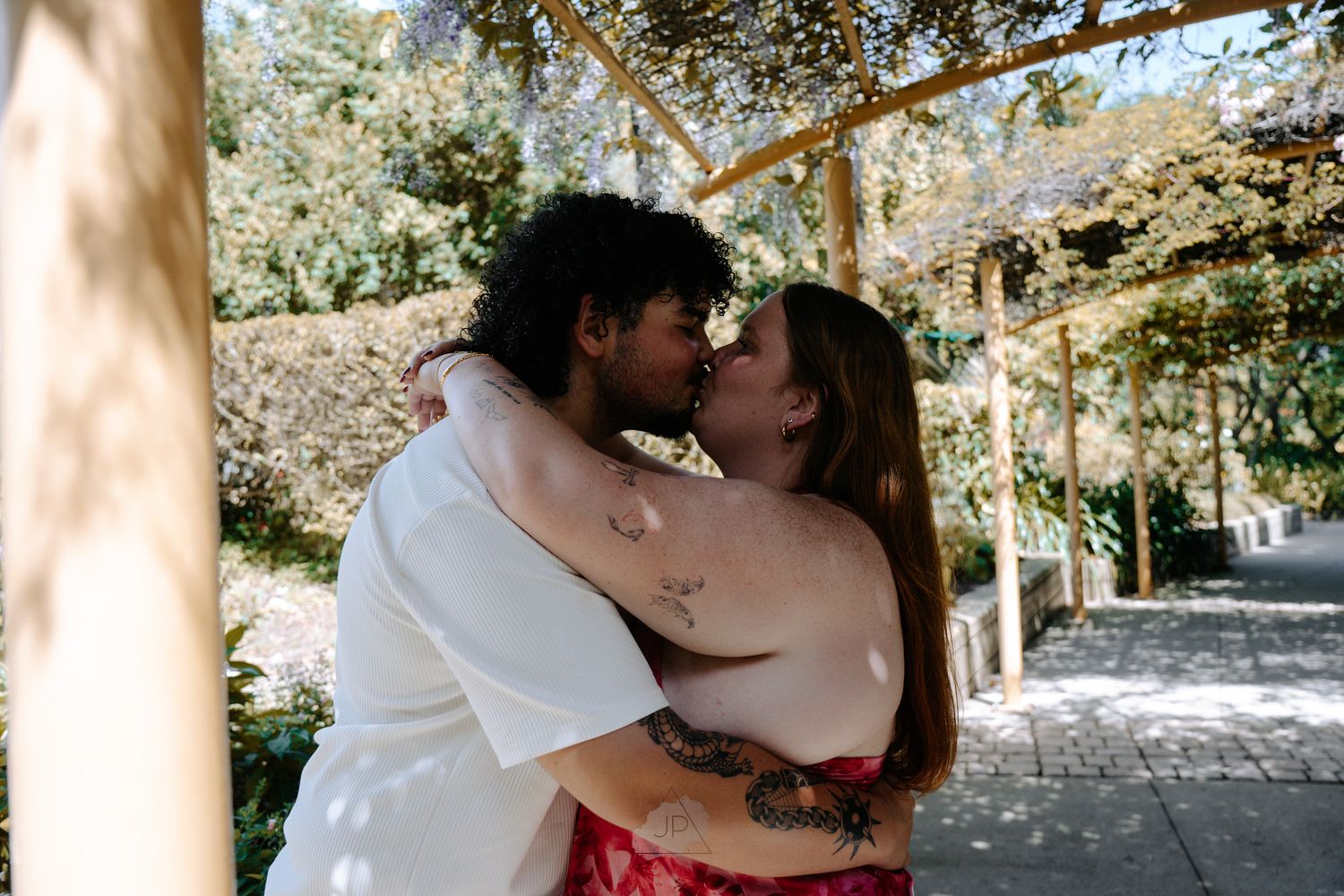 A couple shares an intimate moment beneath a wooden pergola in a garden pathway.