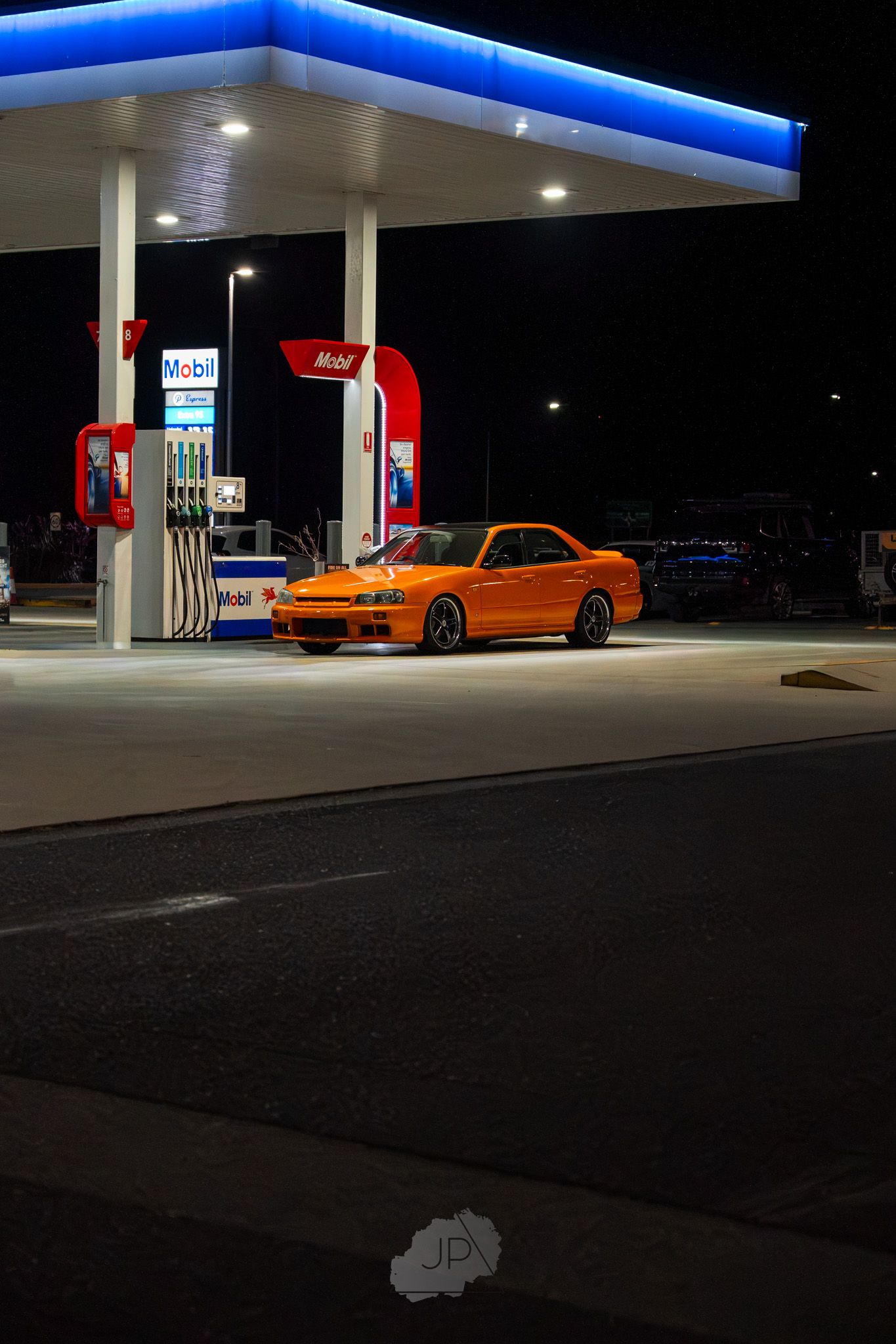 Orange sports car parked at illuminated gas station at night with blue-lit canopy overhead.
