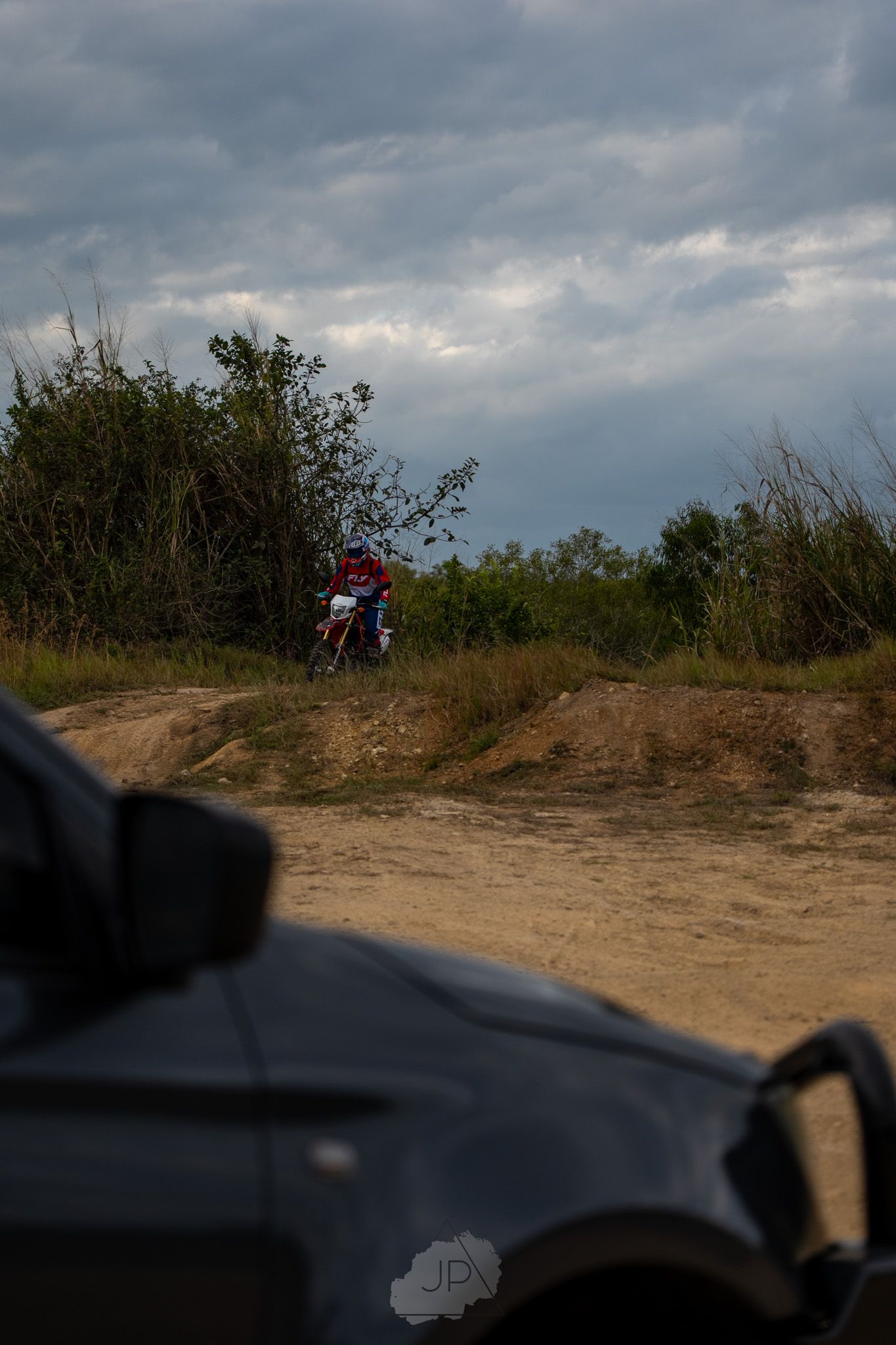 A dirt bike rider navigates a hill on an off-road trail with stormy clouds overhead and a car in the foreground.