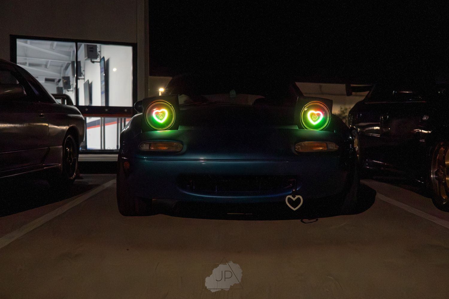 Custom cars parked in a dimly lit lot displaying neon underglow lights in green and blue colors.