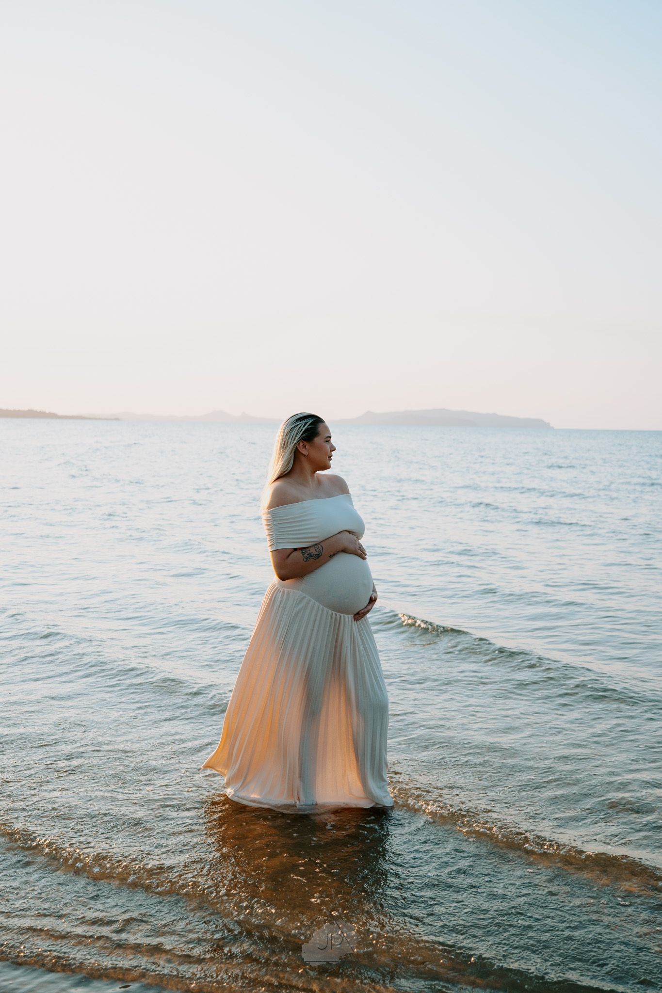 A figure in a flowing turquoise dress stands in shallow ocean waters at sunset creating a dreamy coastal scene.