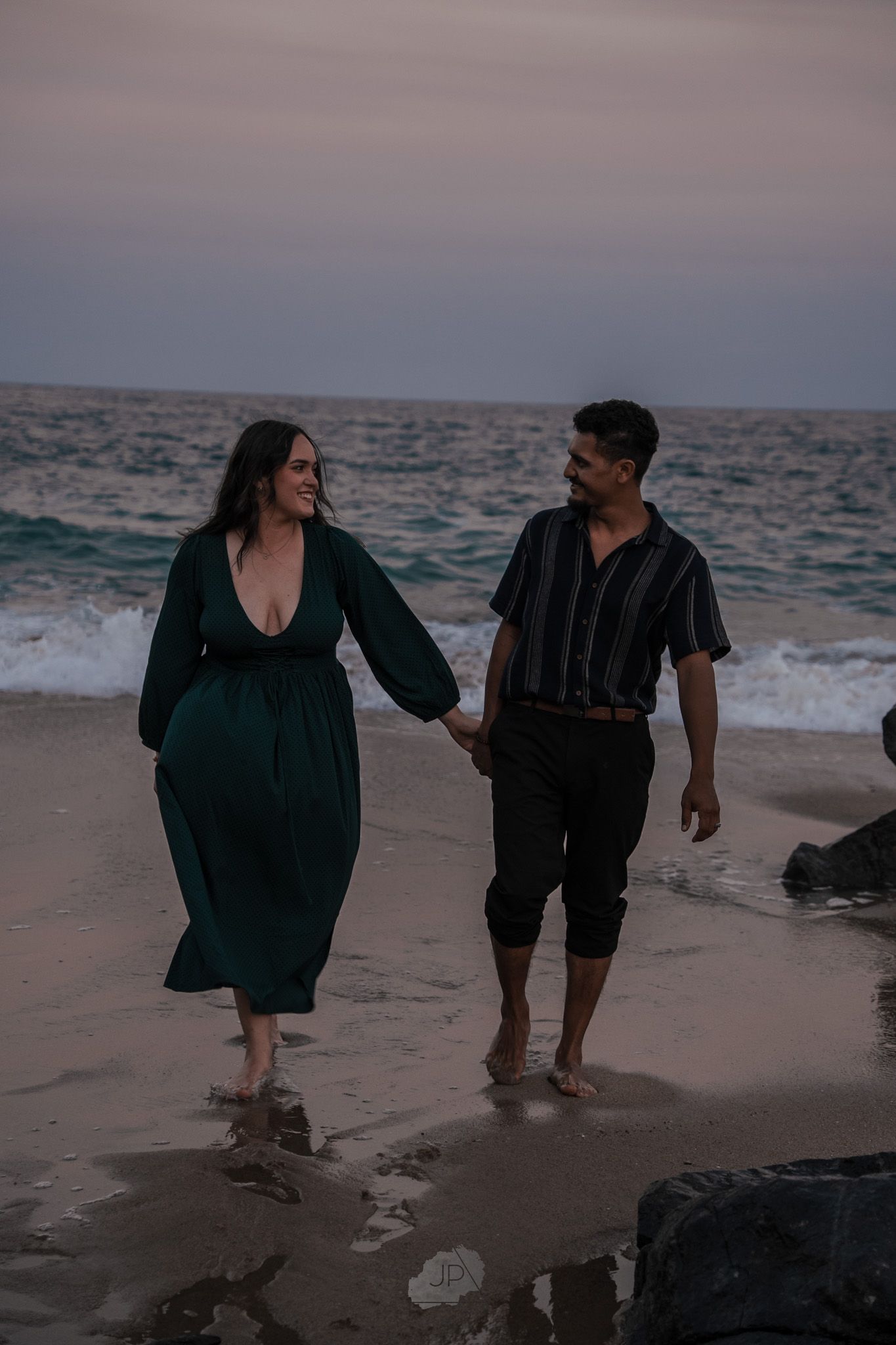 A couple walks hand in hand along a beach at dusk, with waves crashing against dark rocks in the background.