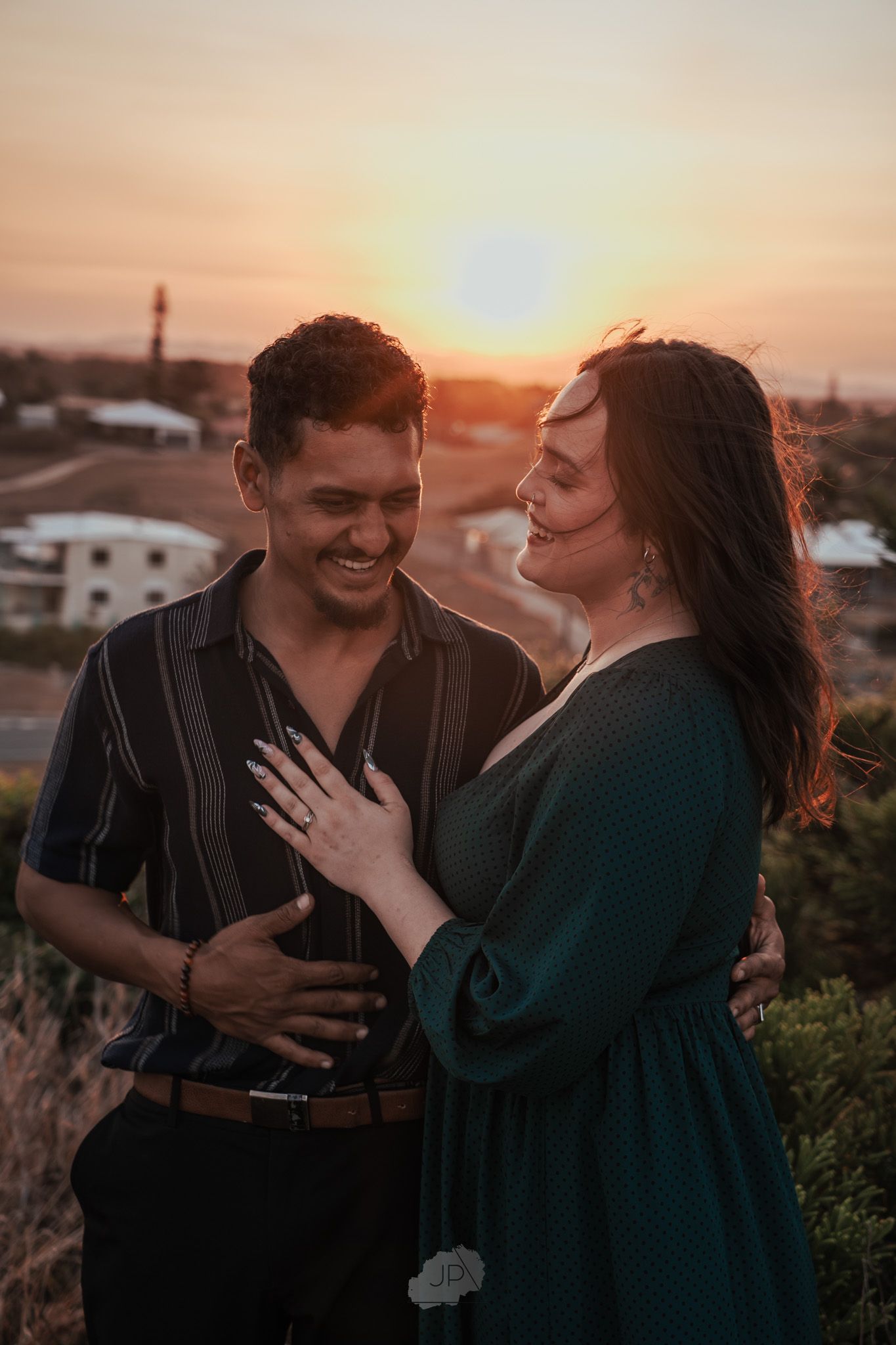 Couple sharing joyful laughter during sunset engagement photoshoot with cityscape in background.