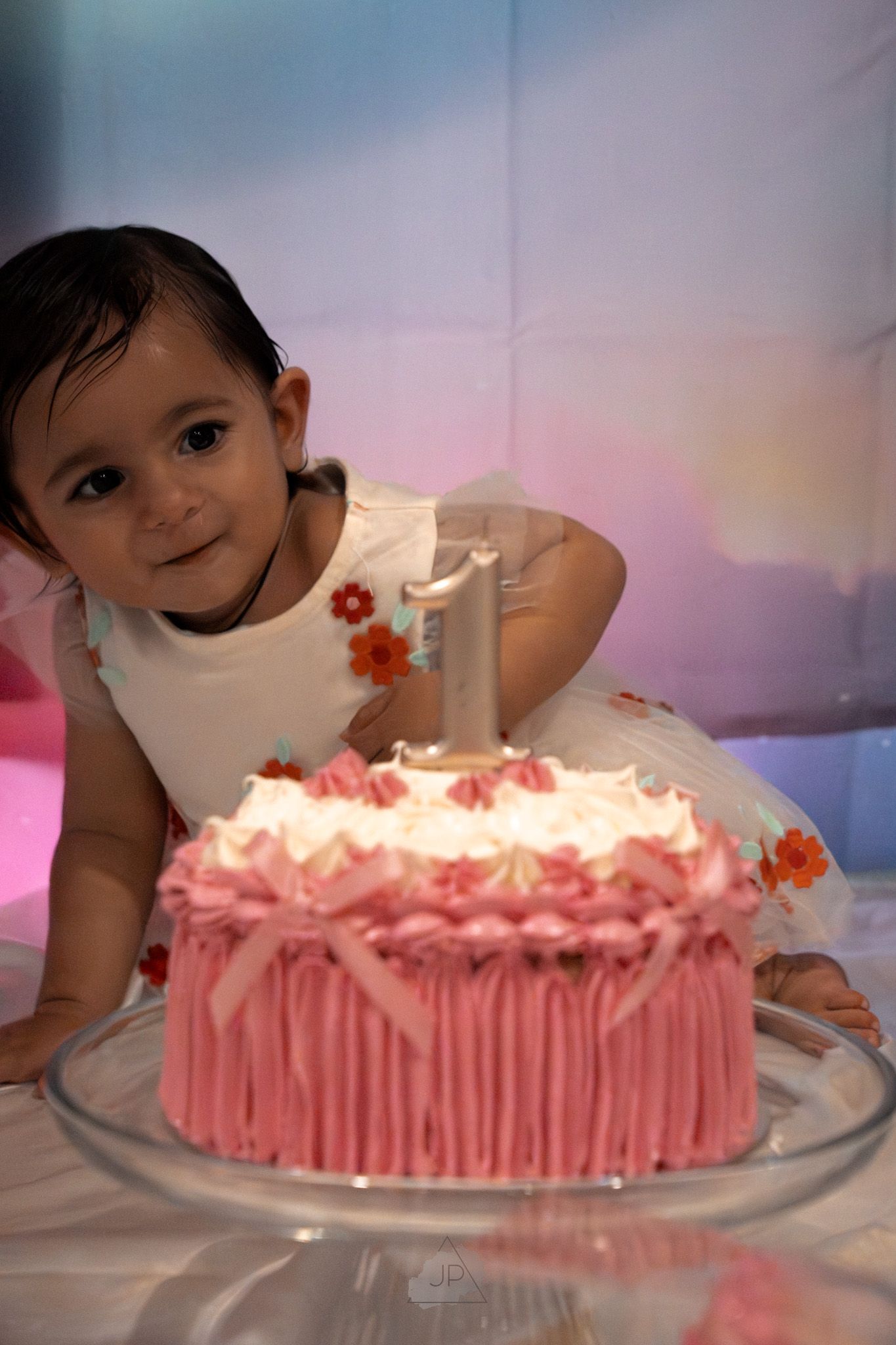 A pink birthday cake with white frosting and a single candle sits on a glass plate at a celebration.