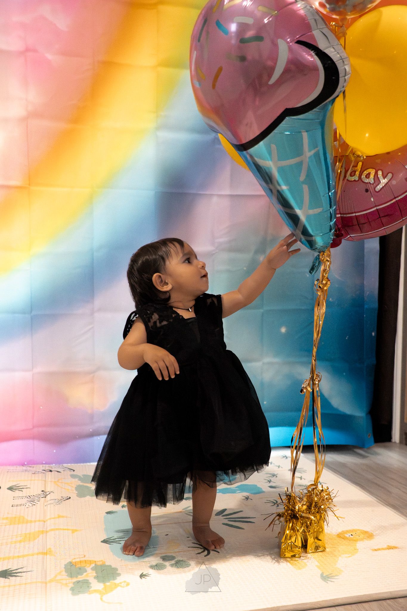 A child in a black tutu dress plays joyfully with colorful balloons against a pastel-colored backdrop during a celebration.