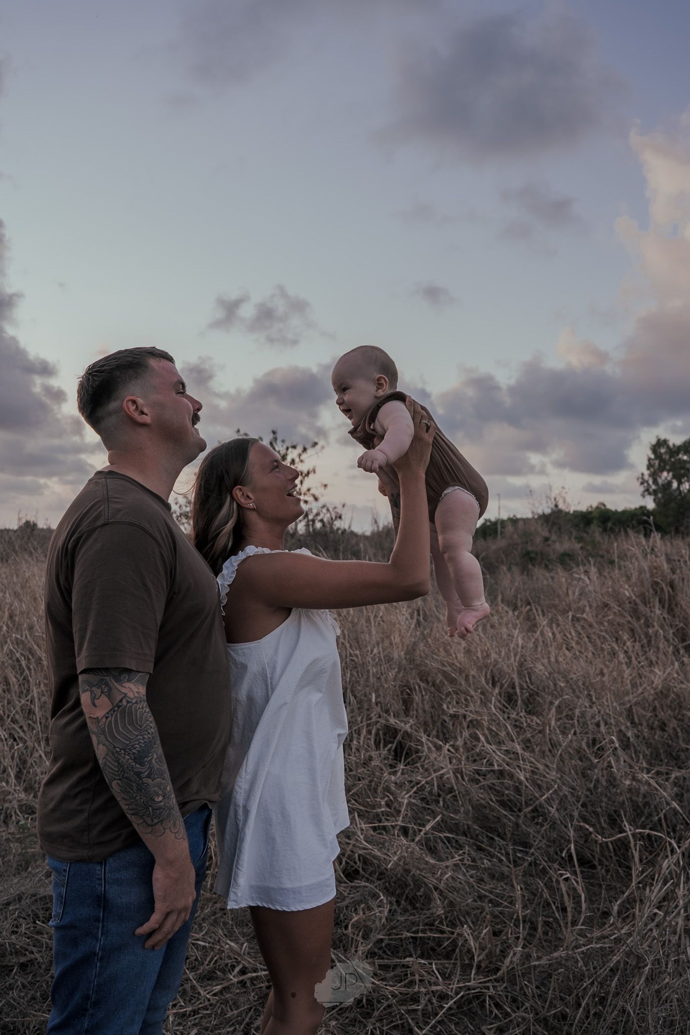 Two people share drinks at sunset in a field of tall grass with dramatic clouds in the background.