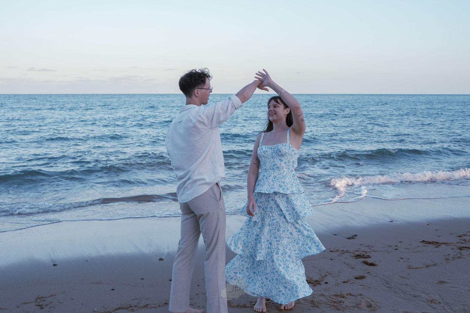 A couple in white and blue outfits dances playfully together on a beach at sunset.