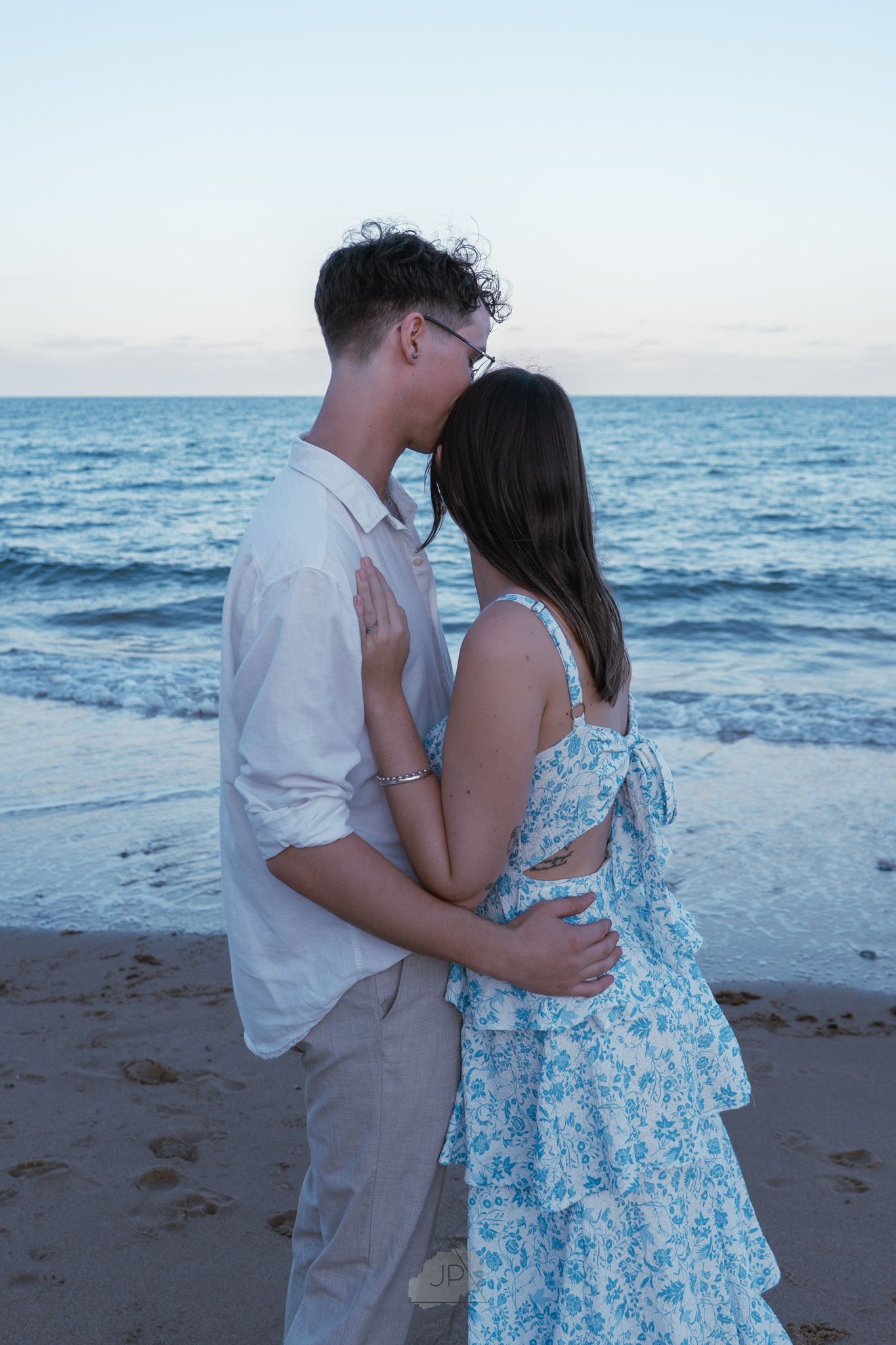 A couple embraces on a beach at sunset wearing white and blue floral outfits near the ocean waves.