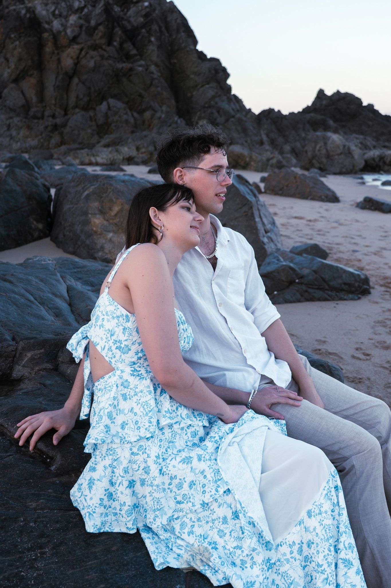 A couple in white and blue floral attire sits together on coastal rocks at sunset, sharing a romantic moment on the beach.