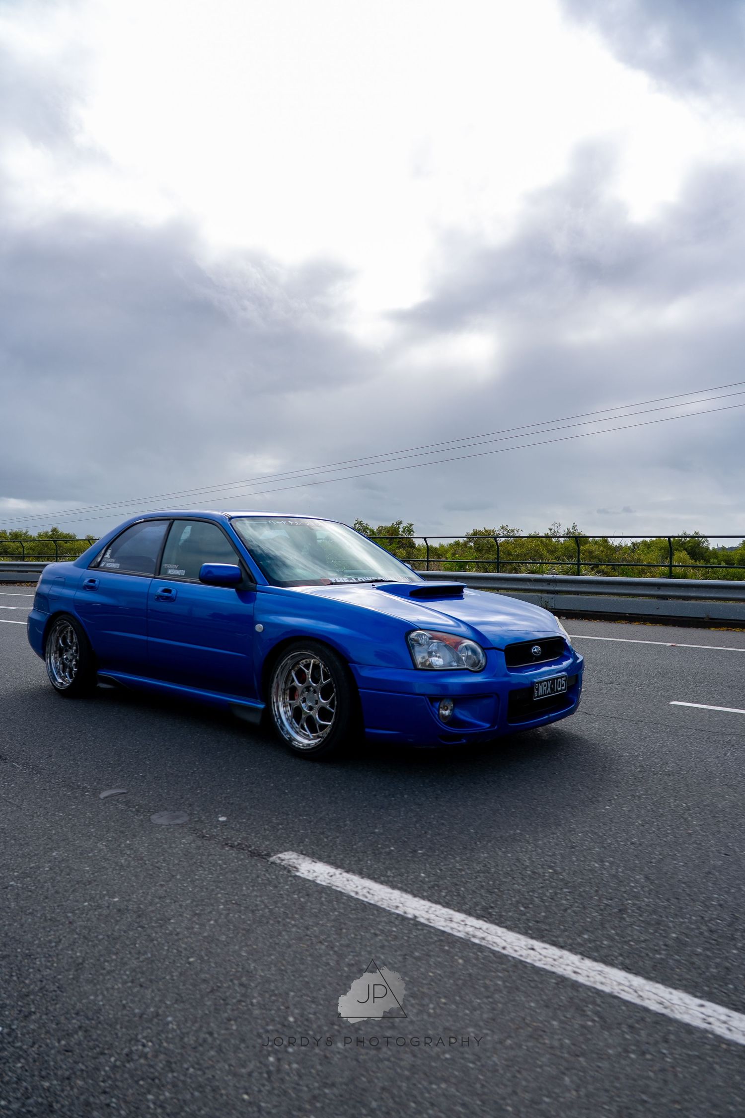 A lowered blue Subaru WRX sits on polished chrome wheels while parked on a highway with cloudy skies overhead.