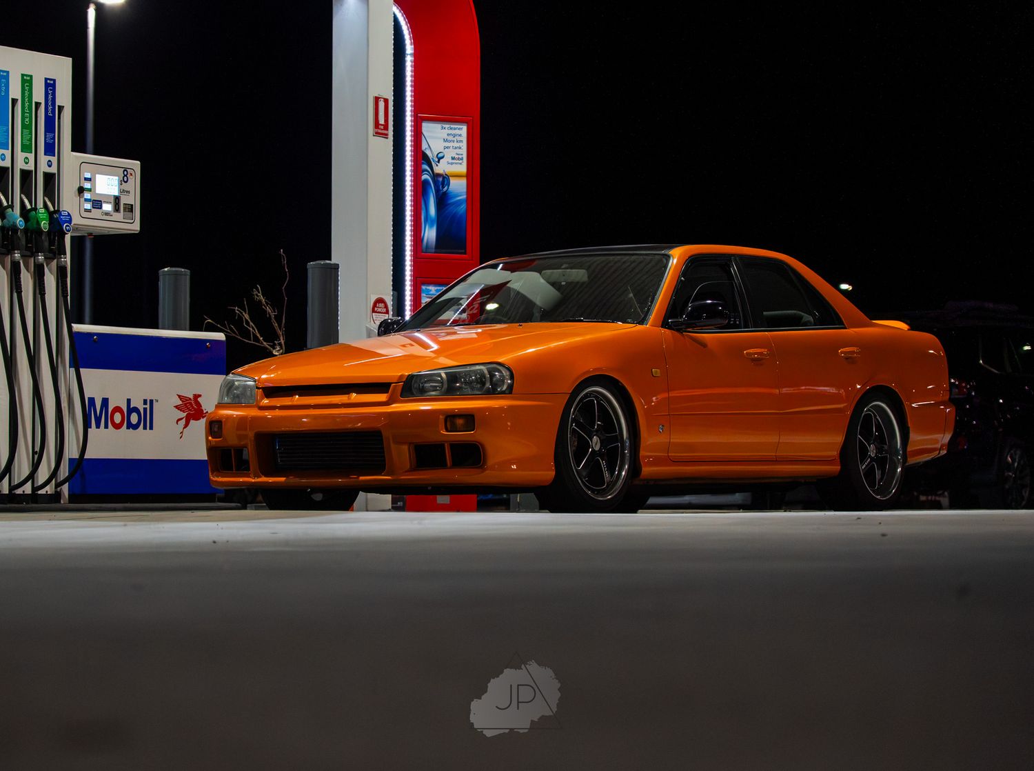 Orange modified Nissan Skyline R33 parked at a gas station at night with Mobil fuel pumps in the background.