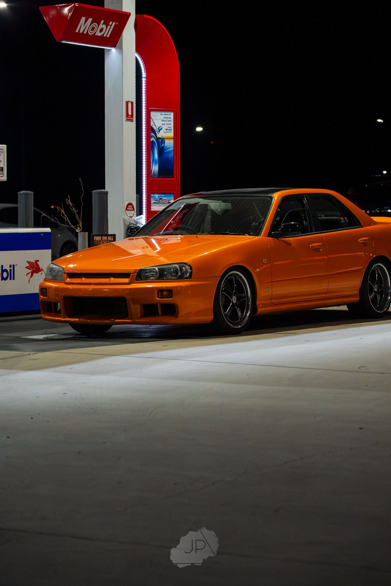 Orange Nissan Skyline R34 GT-R sports car parked at a Mobil gas station at night.