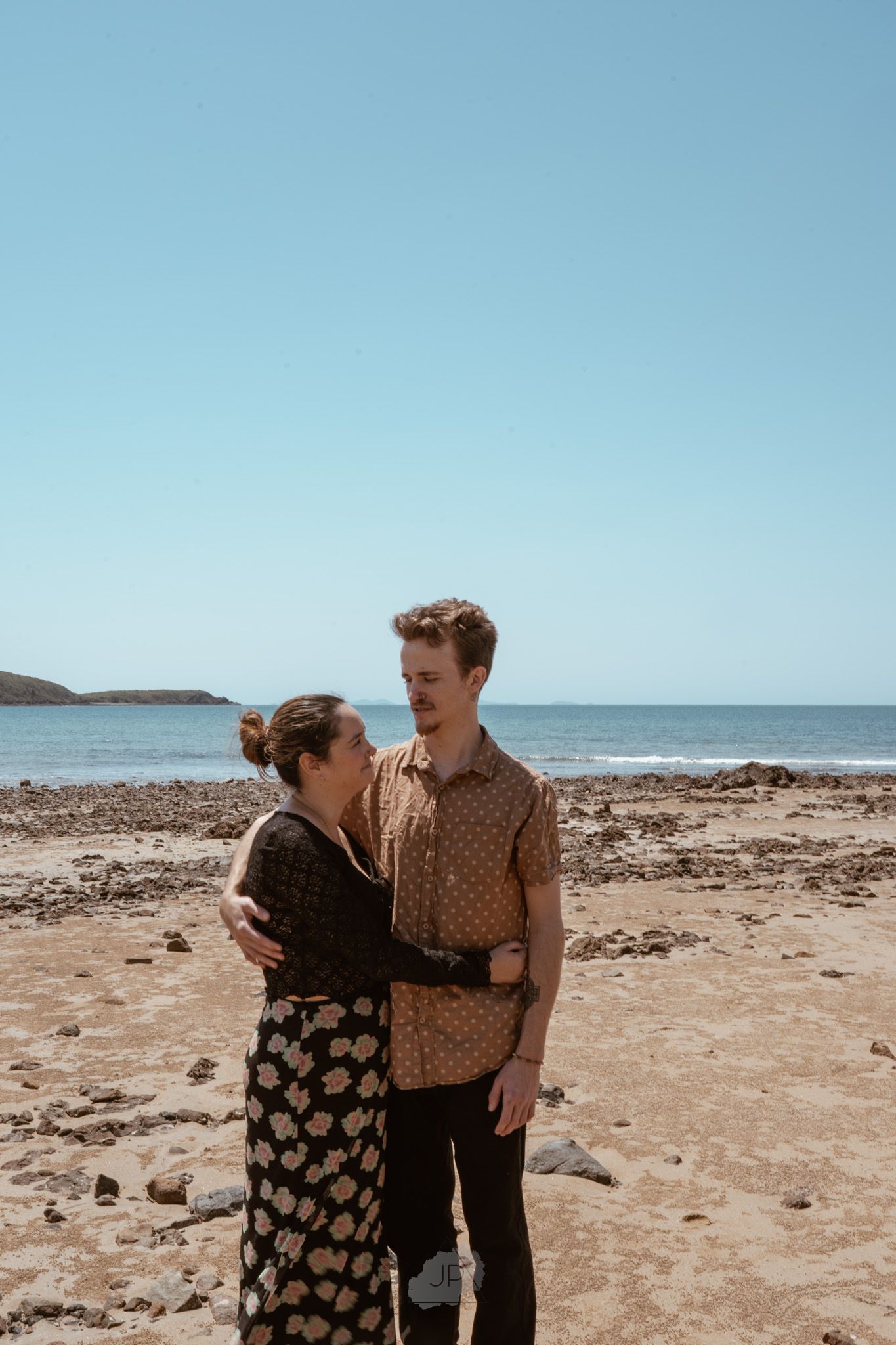 A couple shares a tender moment while walking along a rocky beach shore on a sunny day.