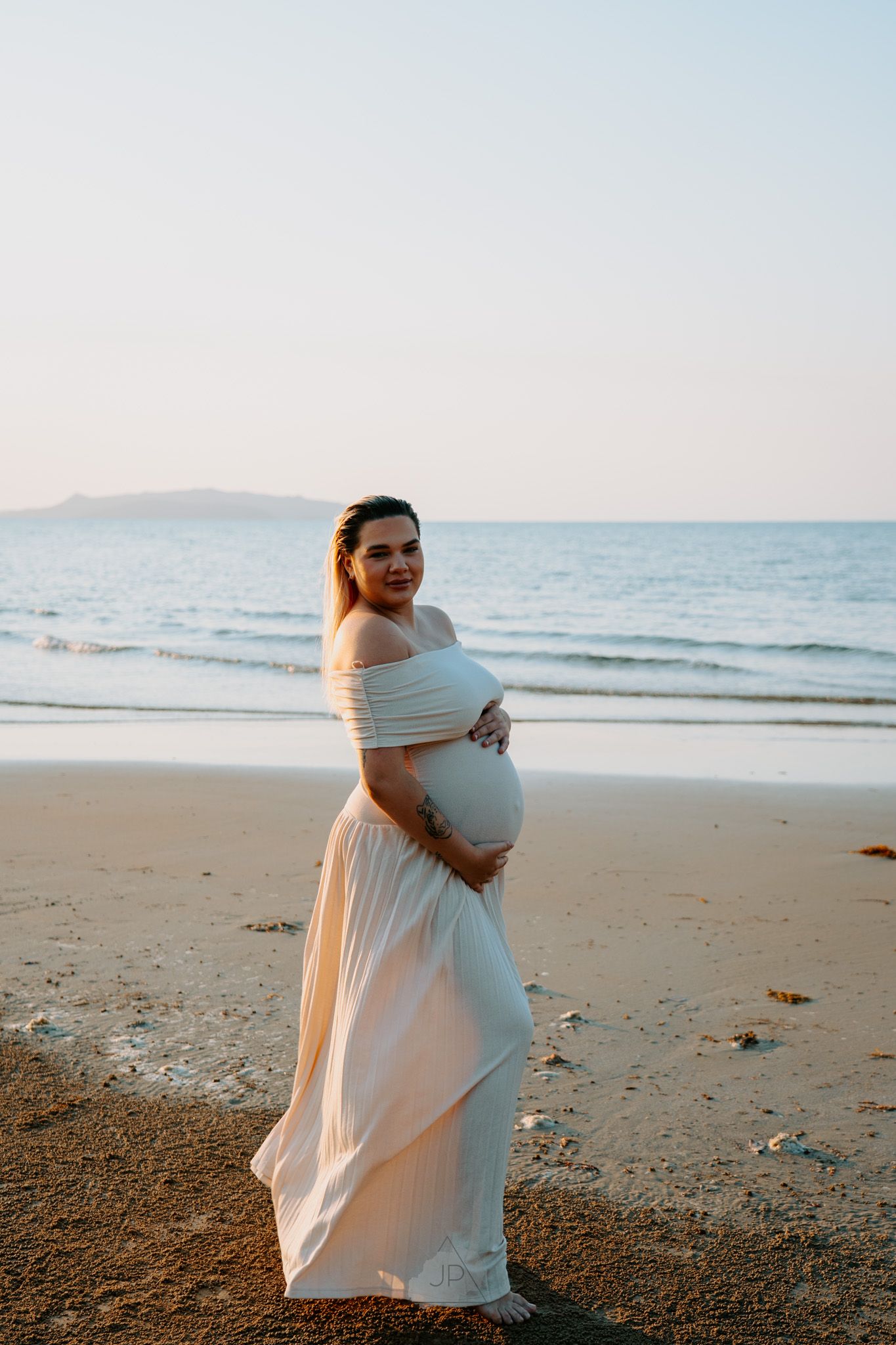 A figure in a flowing white dress poses gracefully against the ocean backdrop during sunset at the beach.