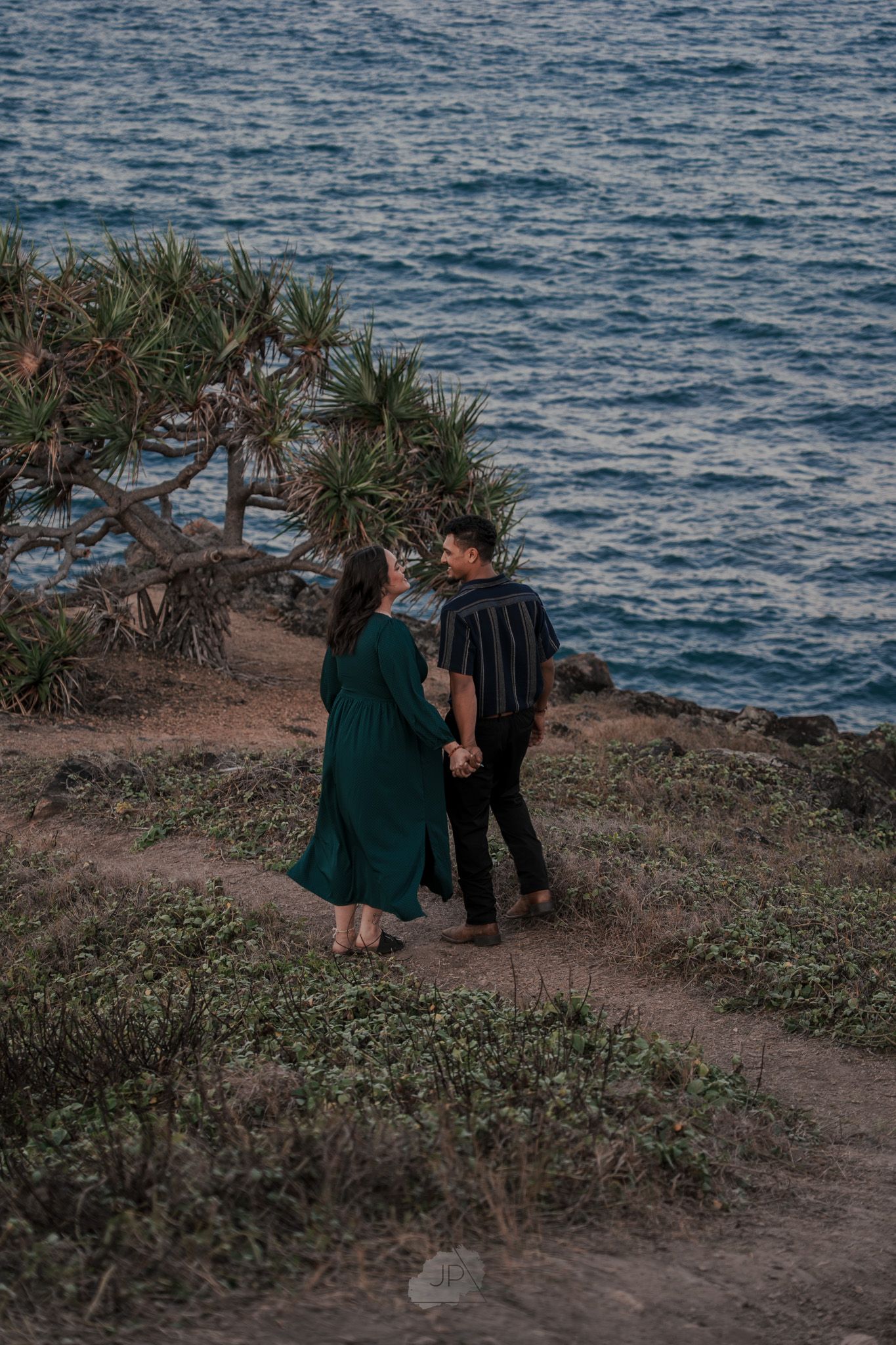 Two people share an intimate moment on a coastal cliff overlooking the ocean at sunset.
