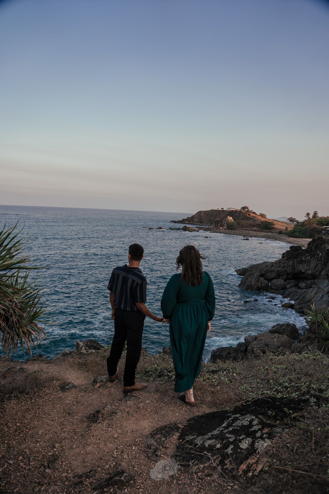 Two people stand together on a rocky coastal cliff overlooking the ocean at sunset.