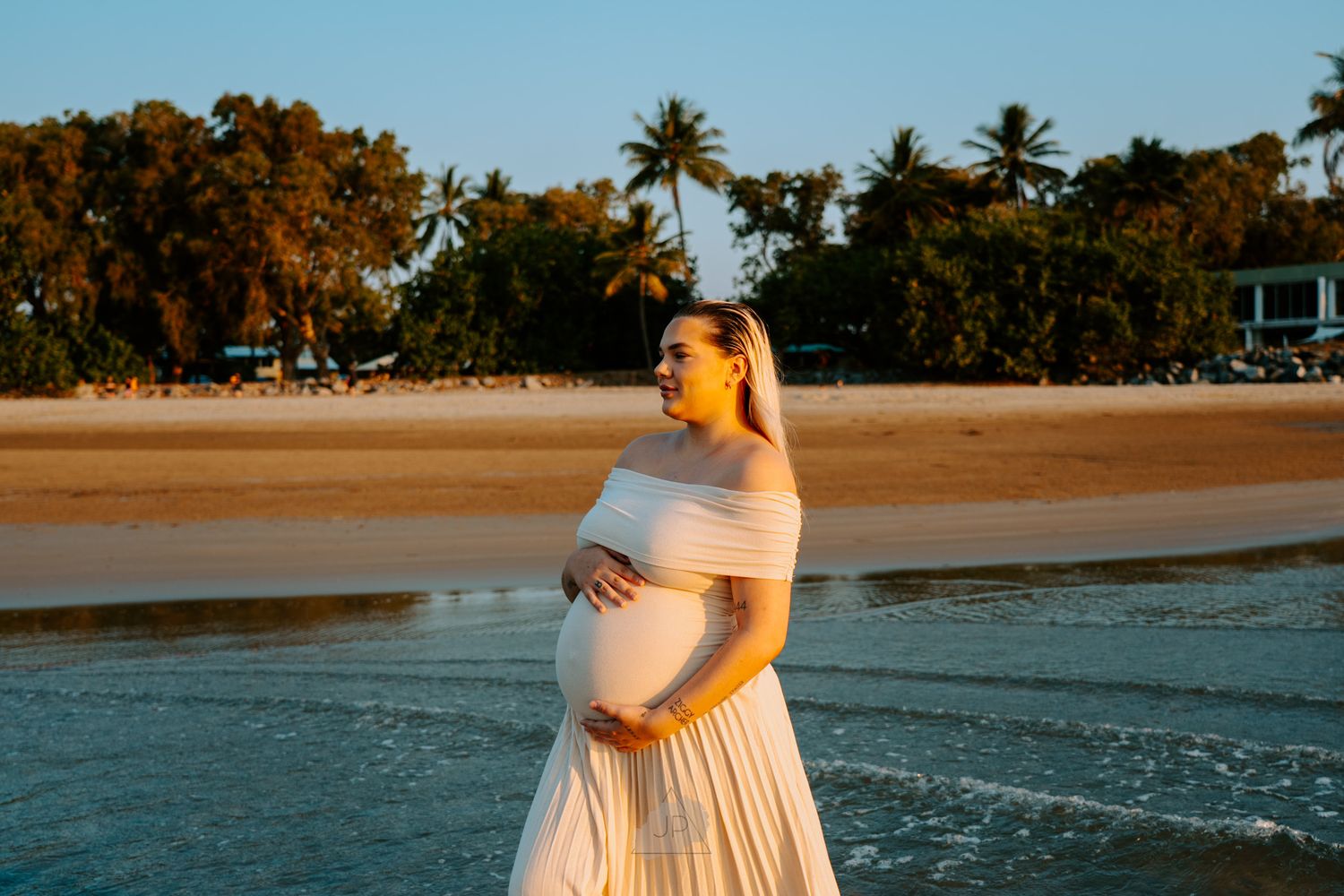 A person in a flowing white dress walks along a tropical beach at sunset with palm trees in the background.