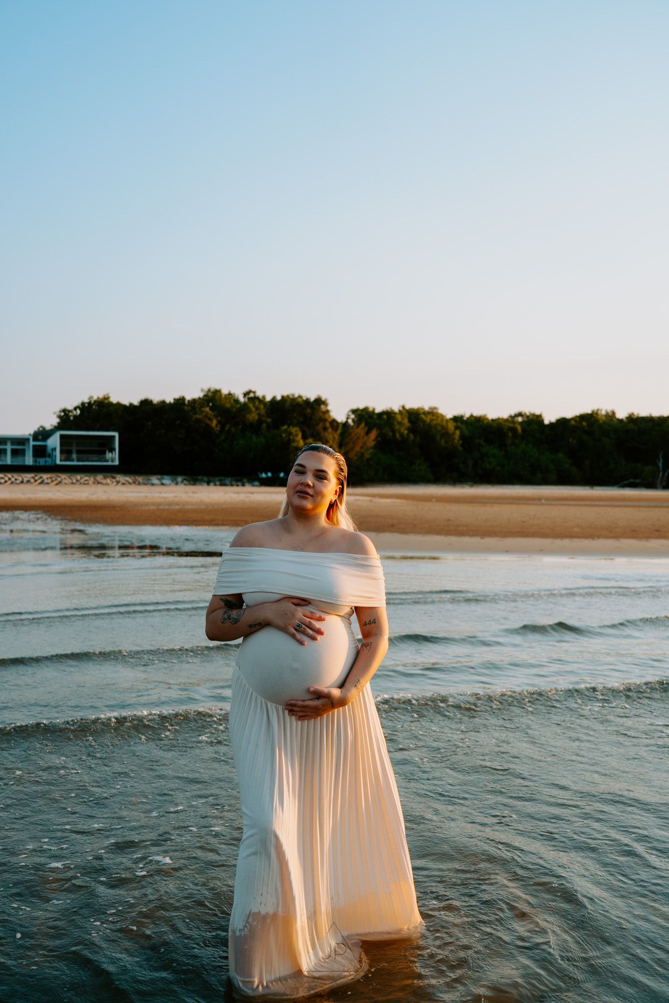 A person in a flowing white dress stands in shallow ocean waves at sunset, creating a dreamy beachside scene.