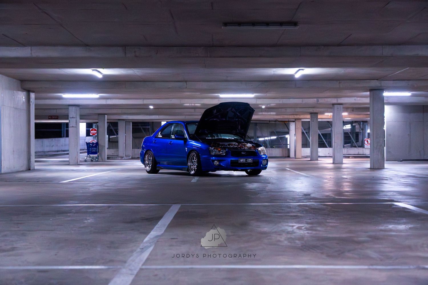 Blue sports car parked in an empty concrete parking garage with fluorescent lighting overhead.