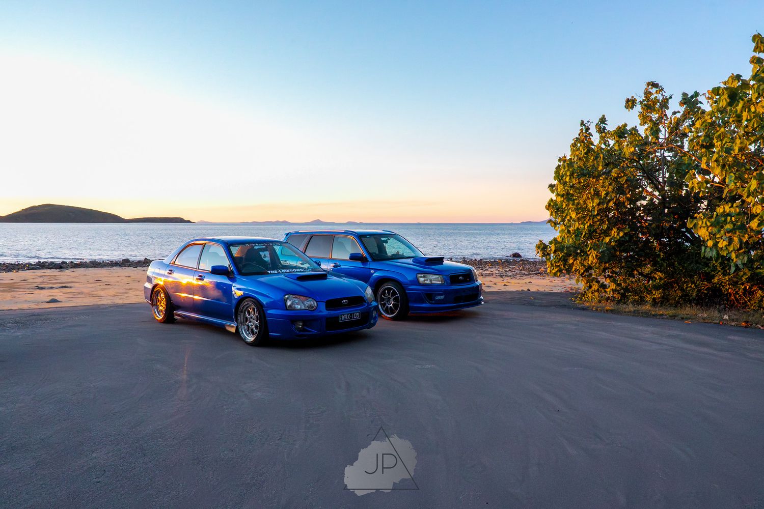 Multiple blue sports cars parked along a waterfront with a sunset view in the background.