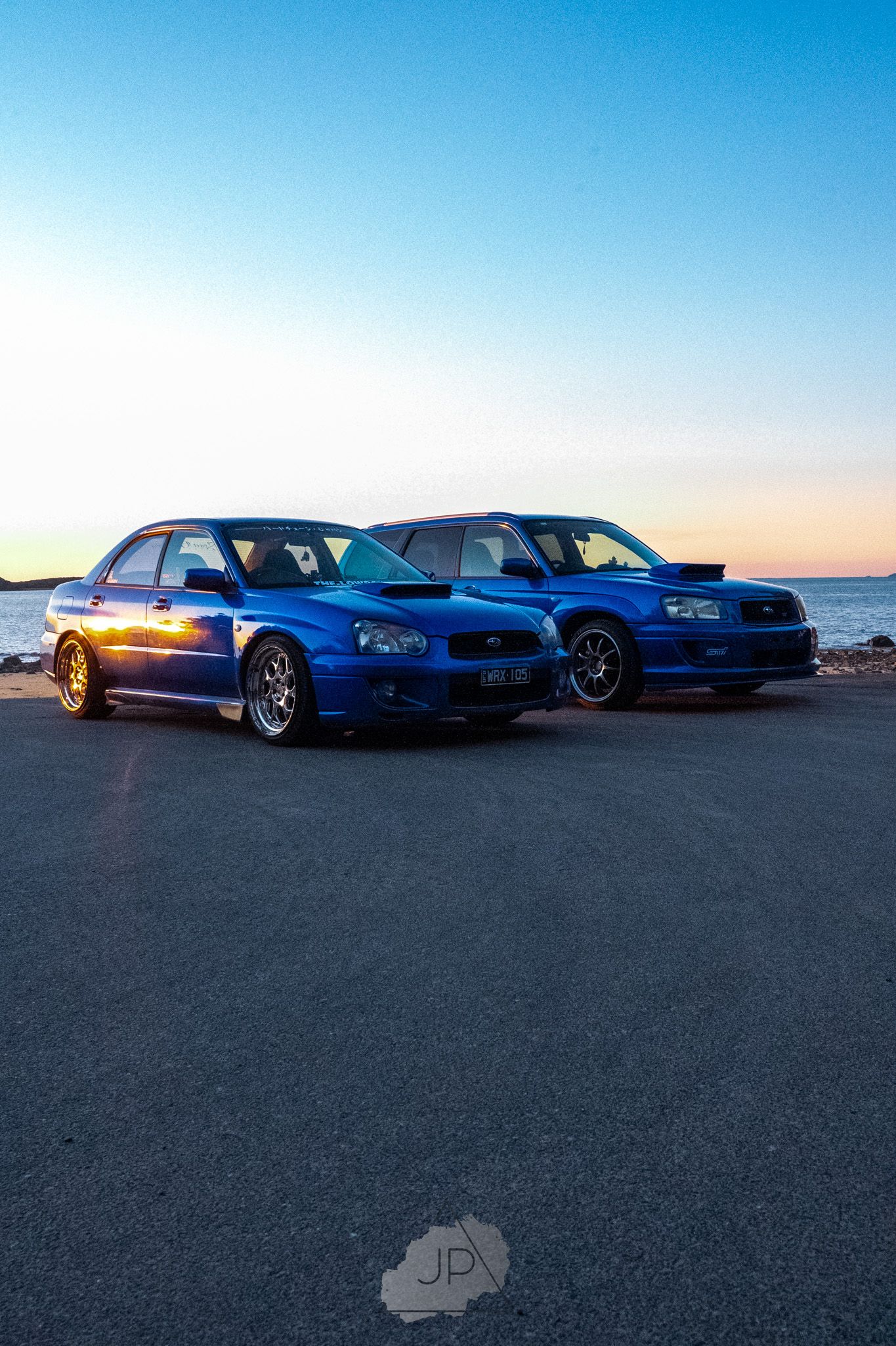 Two blue Subaru WRX sports cars parked on a beach at sunset with vibrant orange sky in the background.