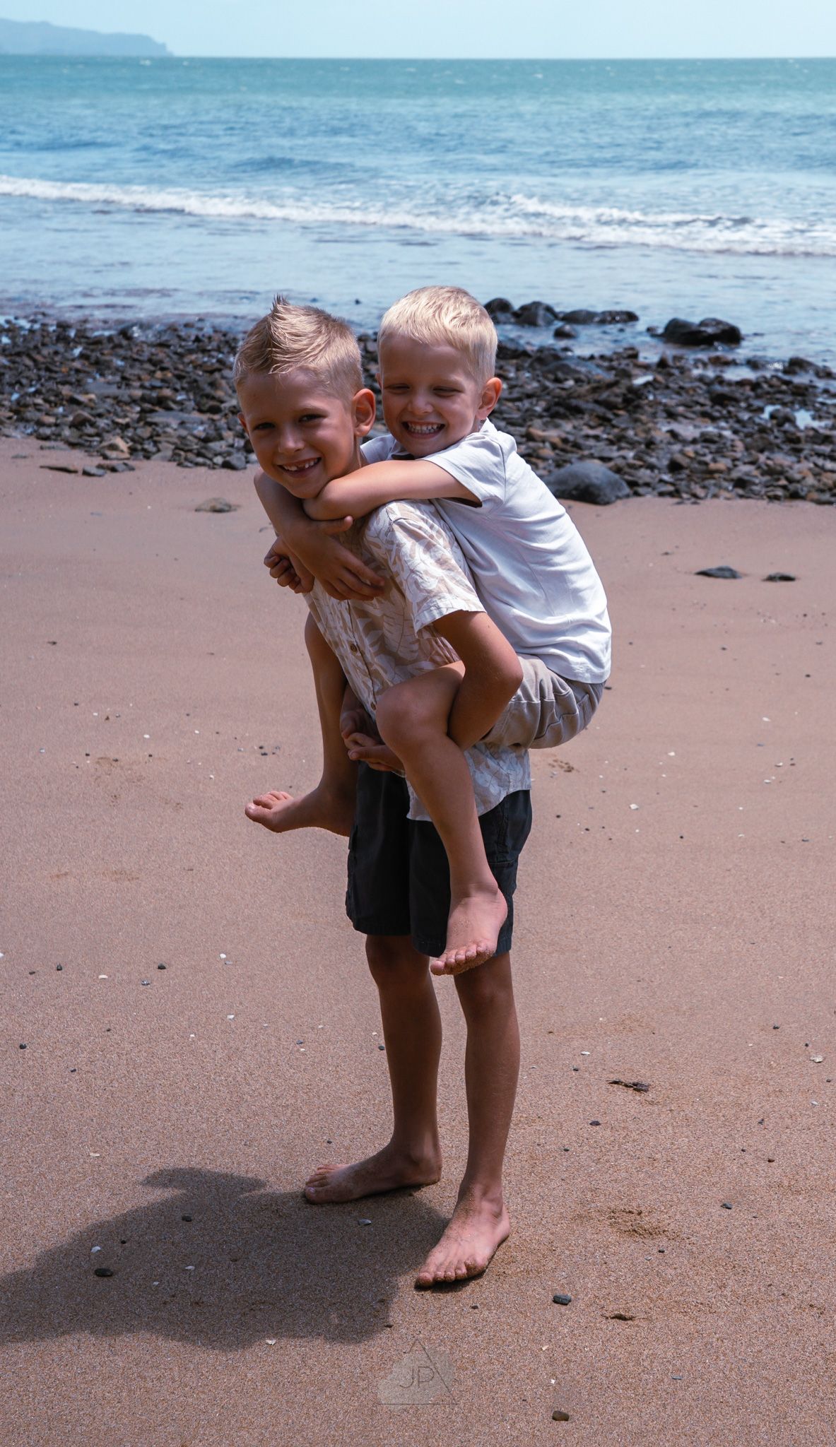 Two young children play together on a sandy beach with rocks and ocean waves in the background.