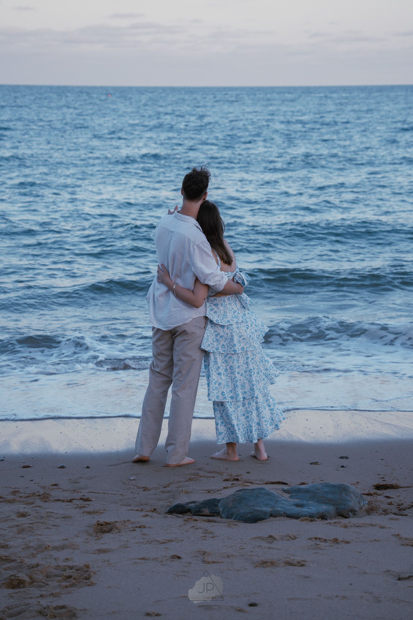 Two people embrace while standing on a sandy beach with the blue ocean waves rolling in behind them.