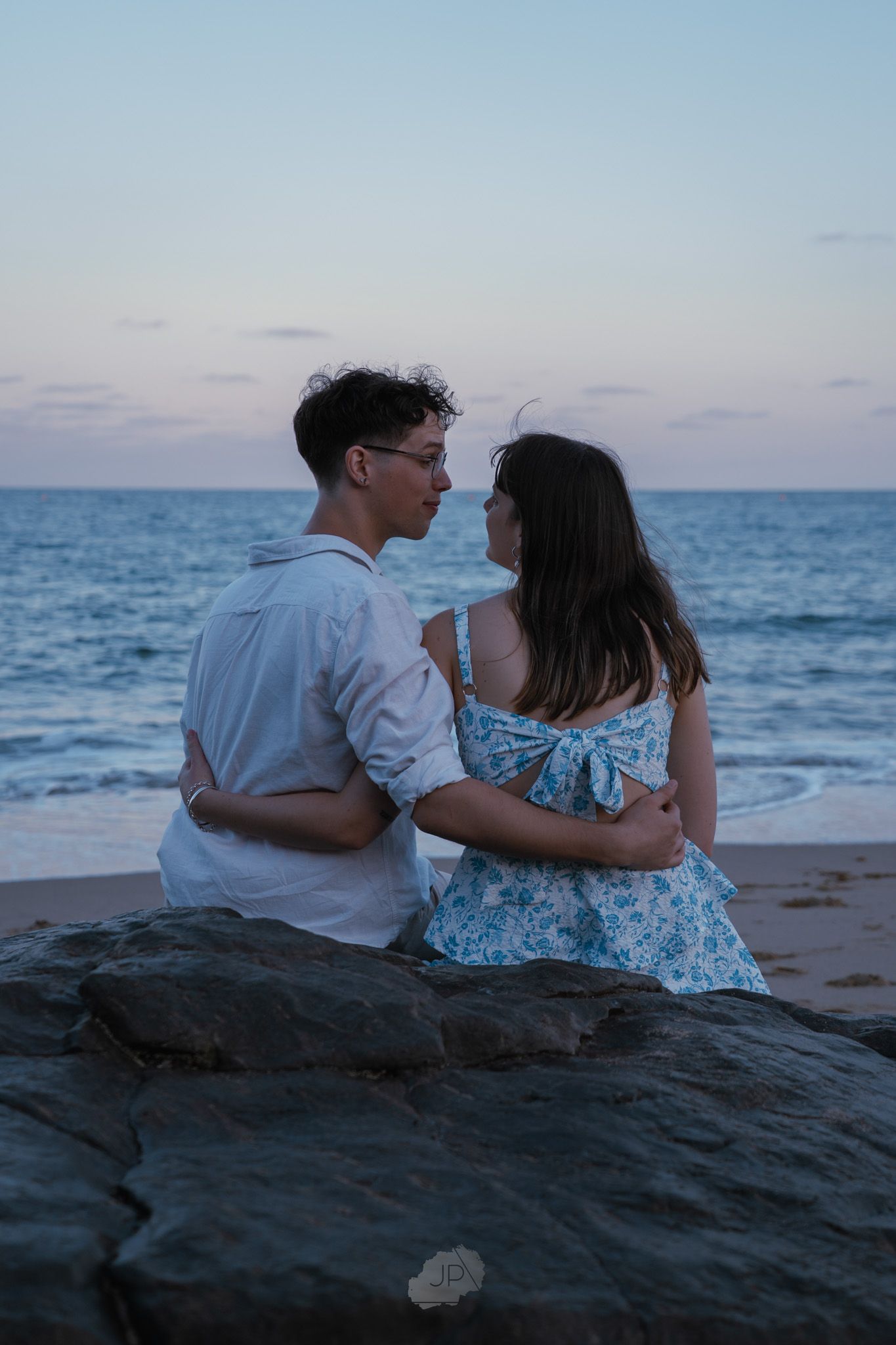 Couple sitting together on rocky beach at sunset watching the ocean waves and horizon.