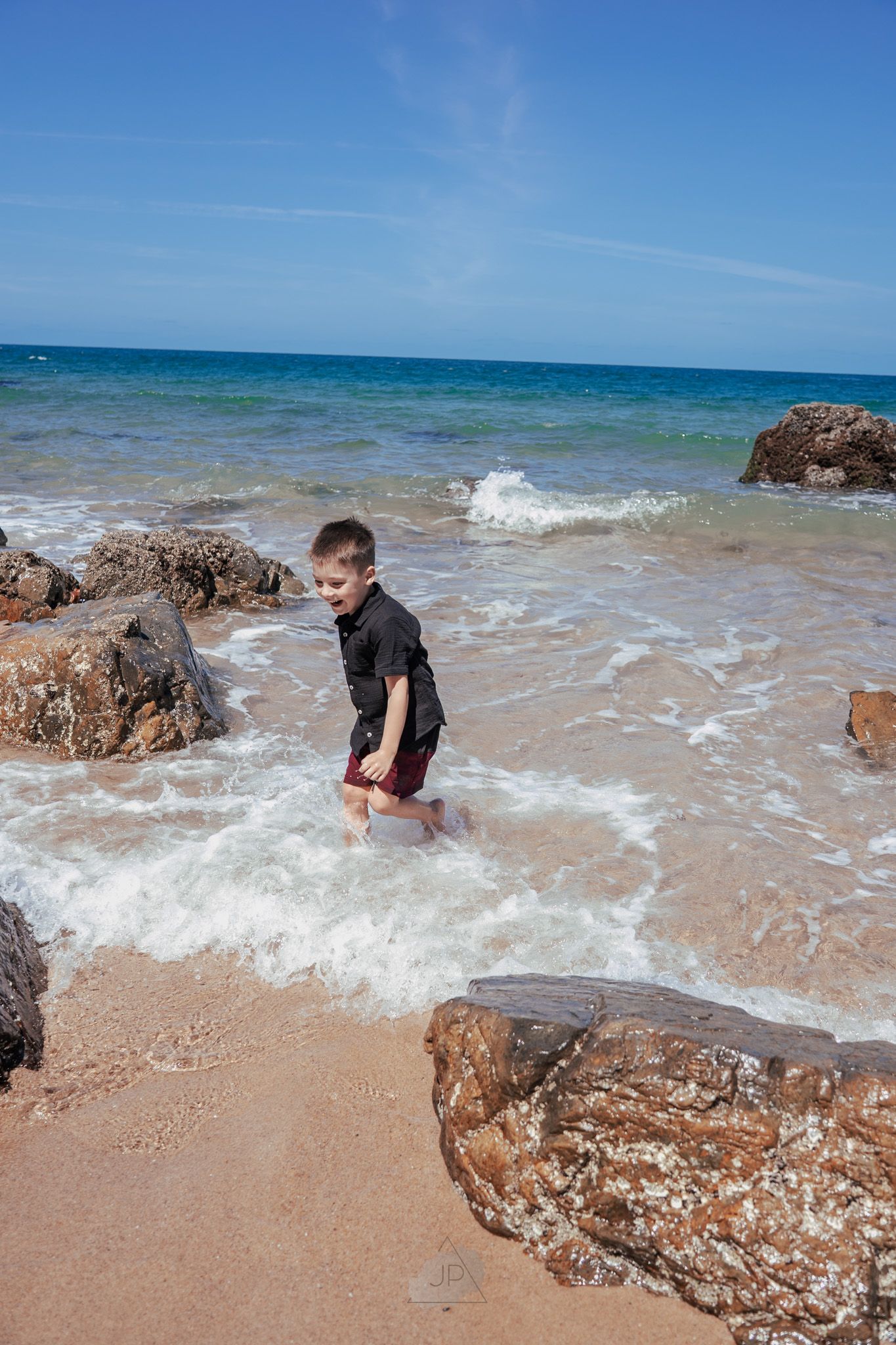 Waves crash against rocky outcrops on a sunny beach with turquoise waters and clear blue skies.