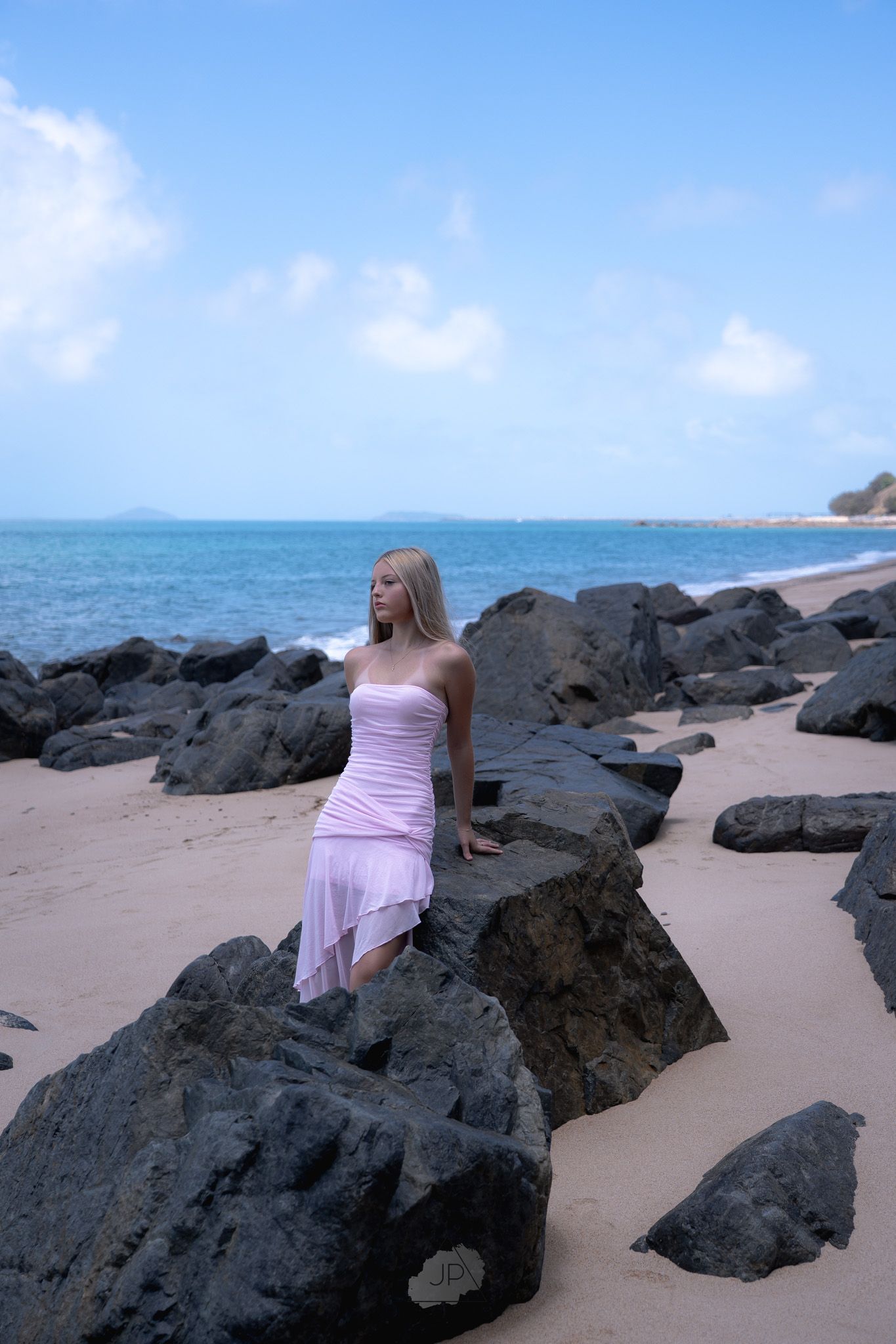 A person in a lavender dress sits among black volcanic rocks on a tropical beach with ocean views.