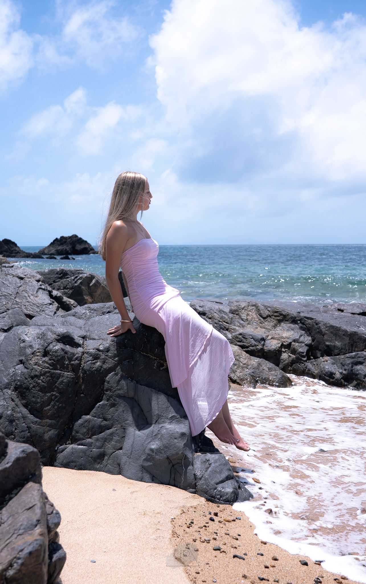 A person in a pink dress sits on dark volcanic rocks along a tropical coastline with blue ocean and sky in the background.